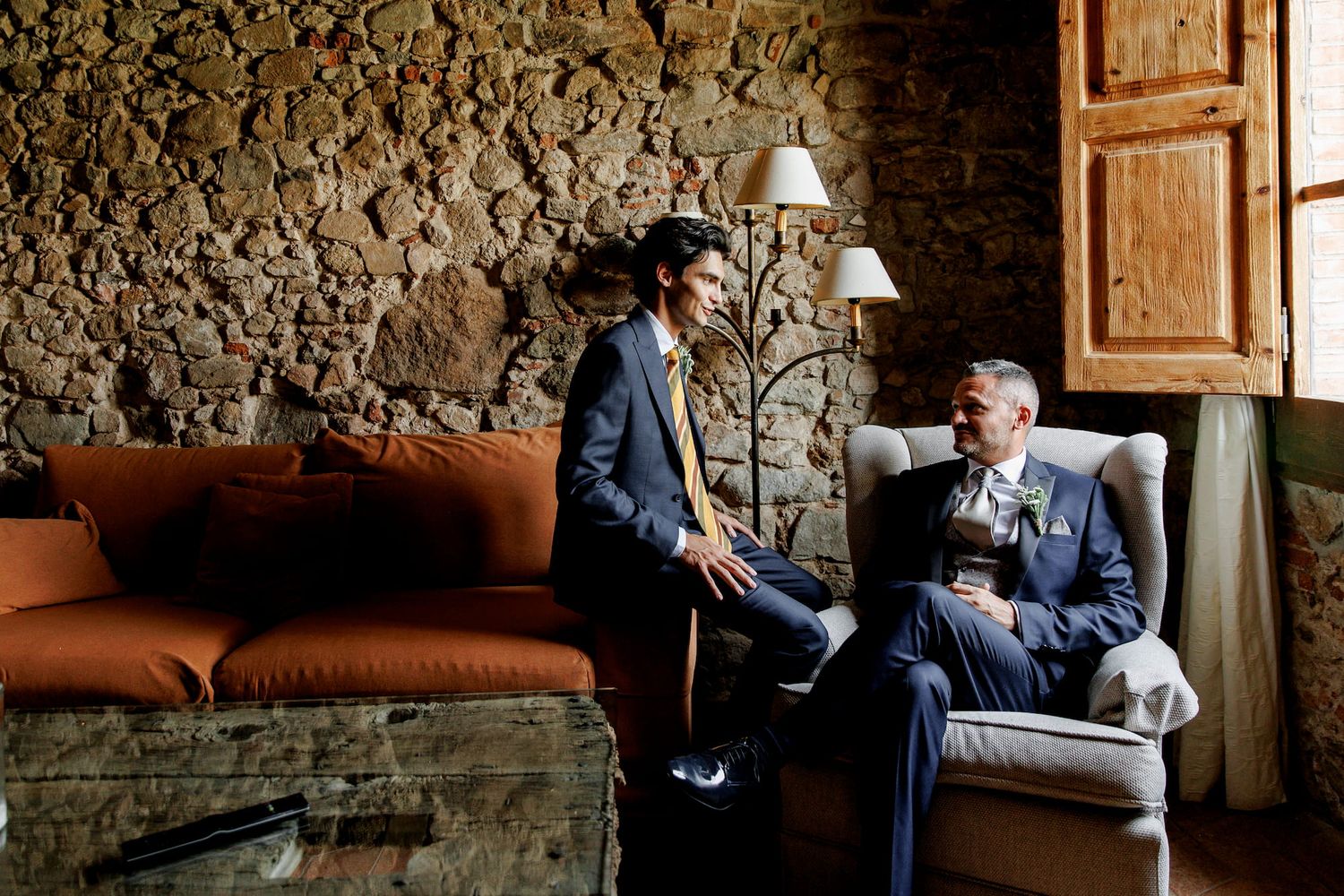 Two grooms in suits share a conversation in a rustic stone-walled venue before their wedding ceremony.