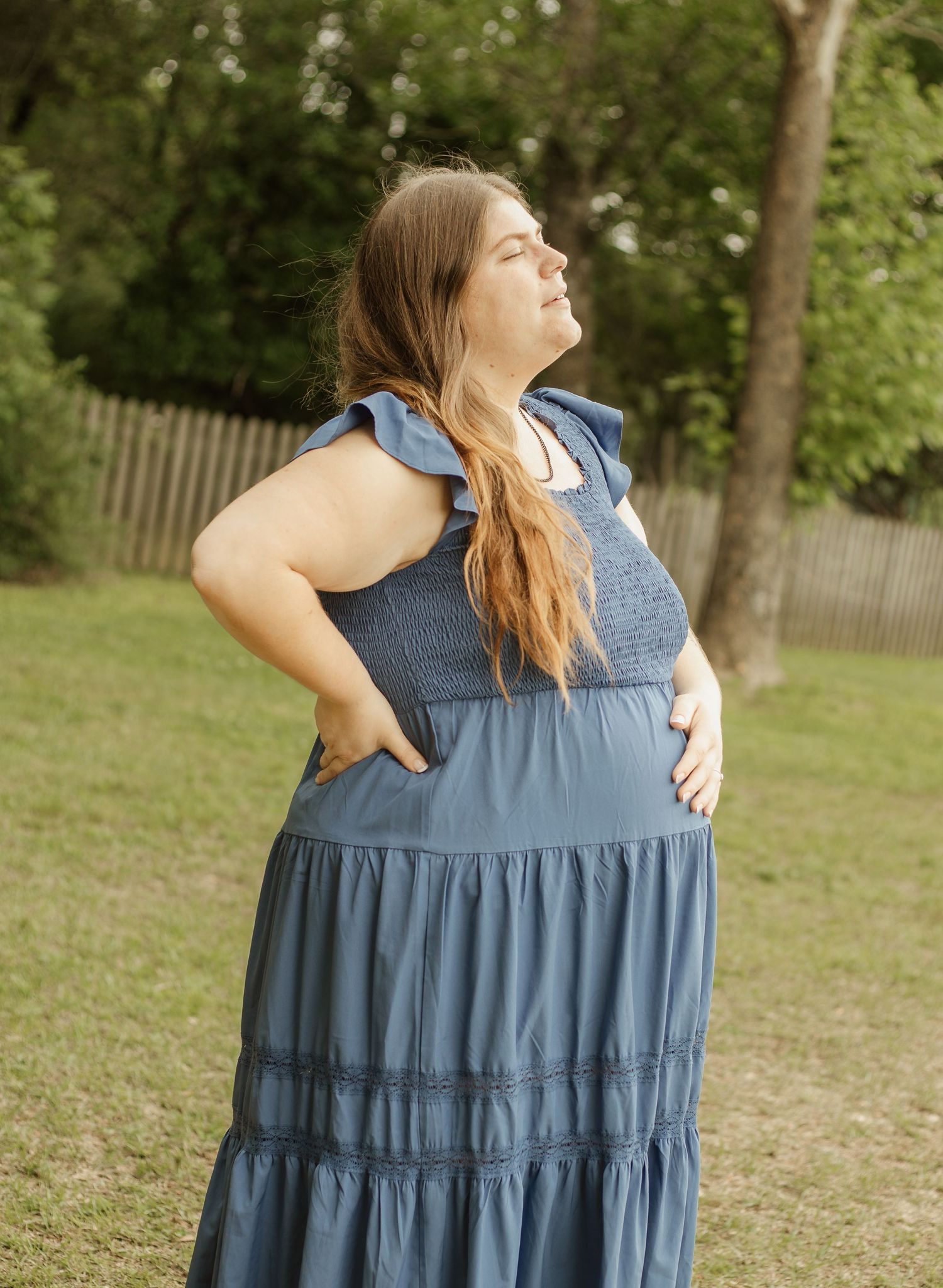 Woman in blue tiered maxi dress posing outdoors in a backyard setting with trees and wooden fence.