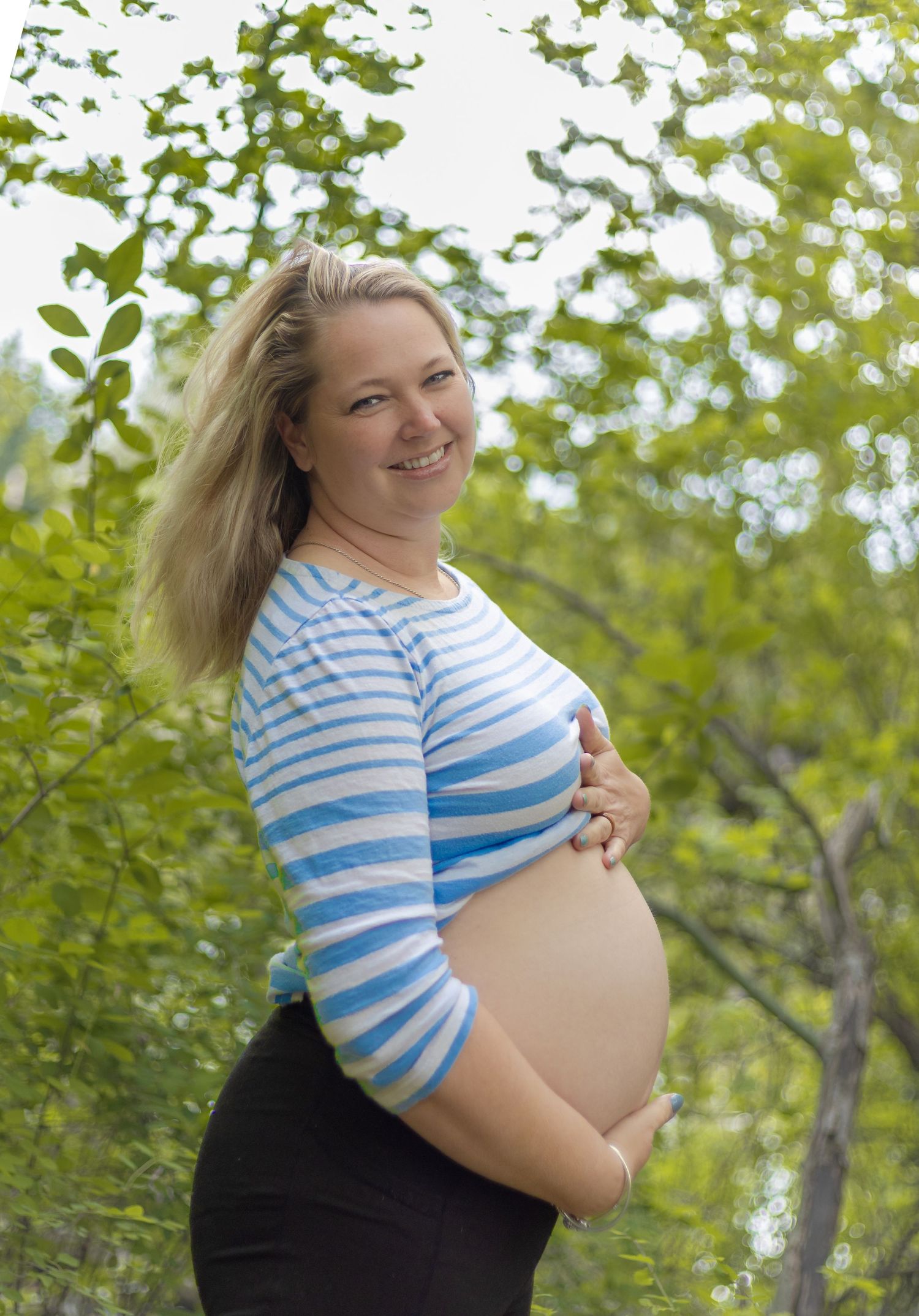 Smiling pregnant woman in striped top cradling baby bump outdoors among green trees.