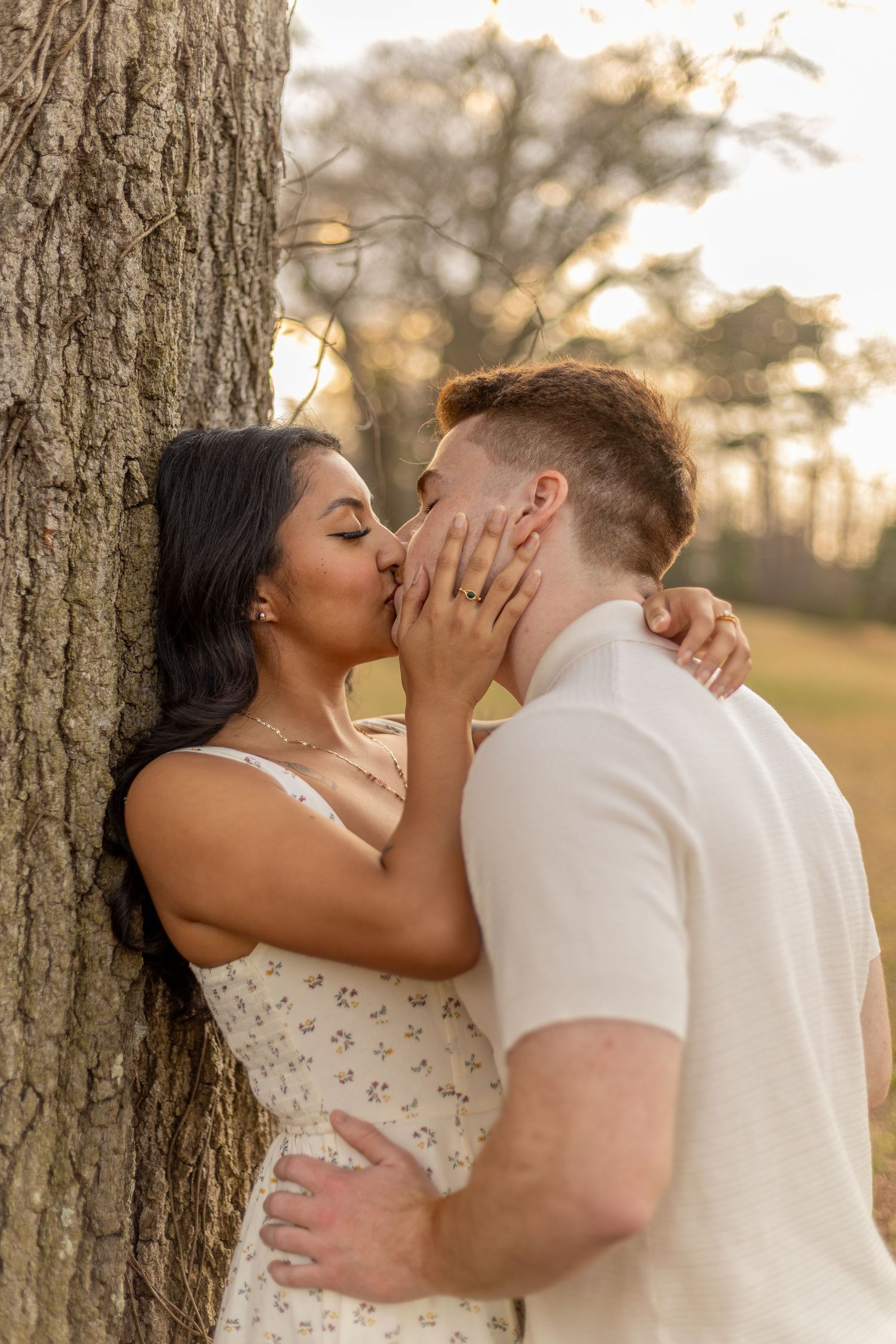 Intimate couple sharing a passionate kiss against a tree trunk at sunset.