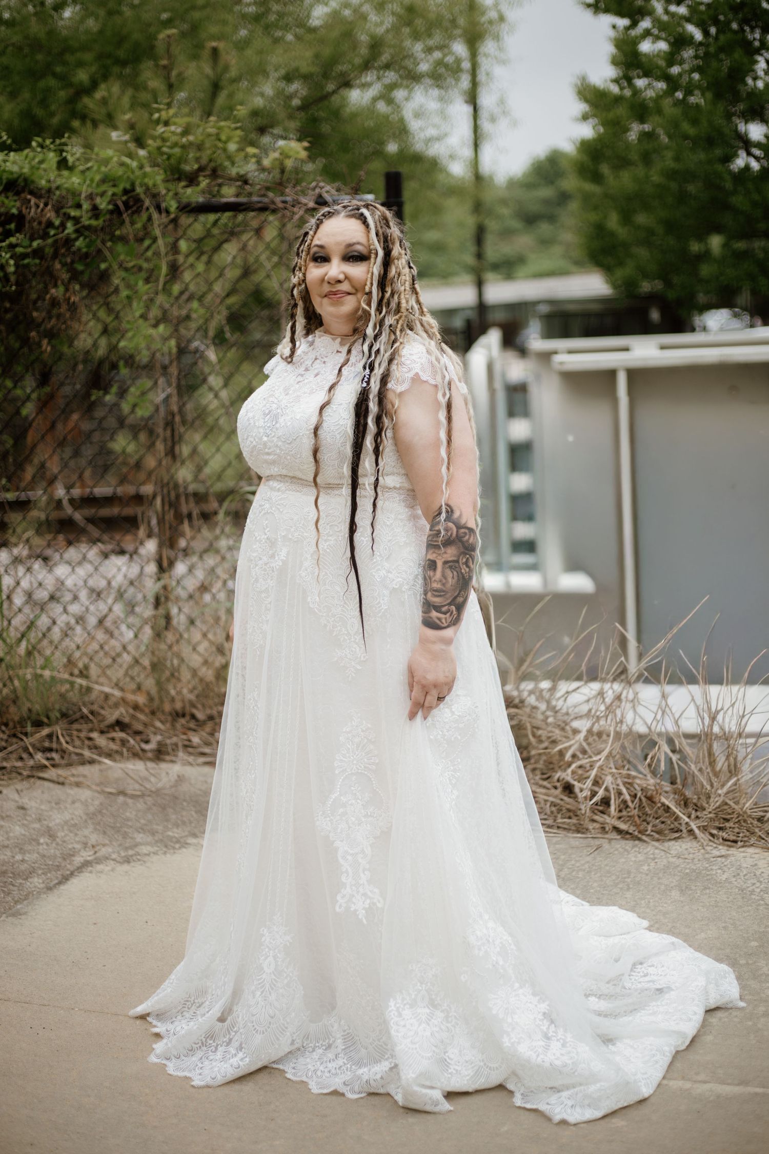 Plus-size bride in white lace wedding gown with long train, colorful braids, and sleeve tattoos posing outdoors.