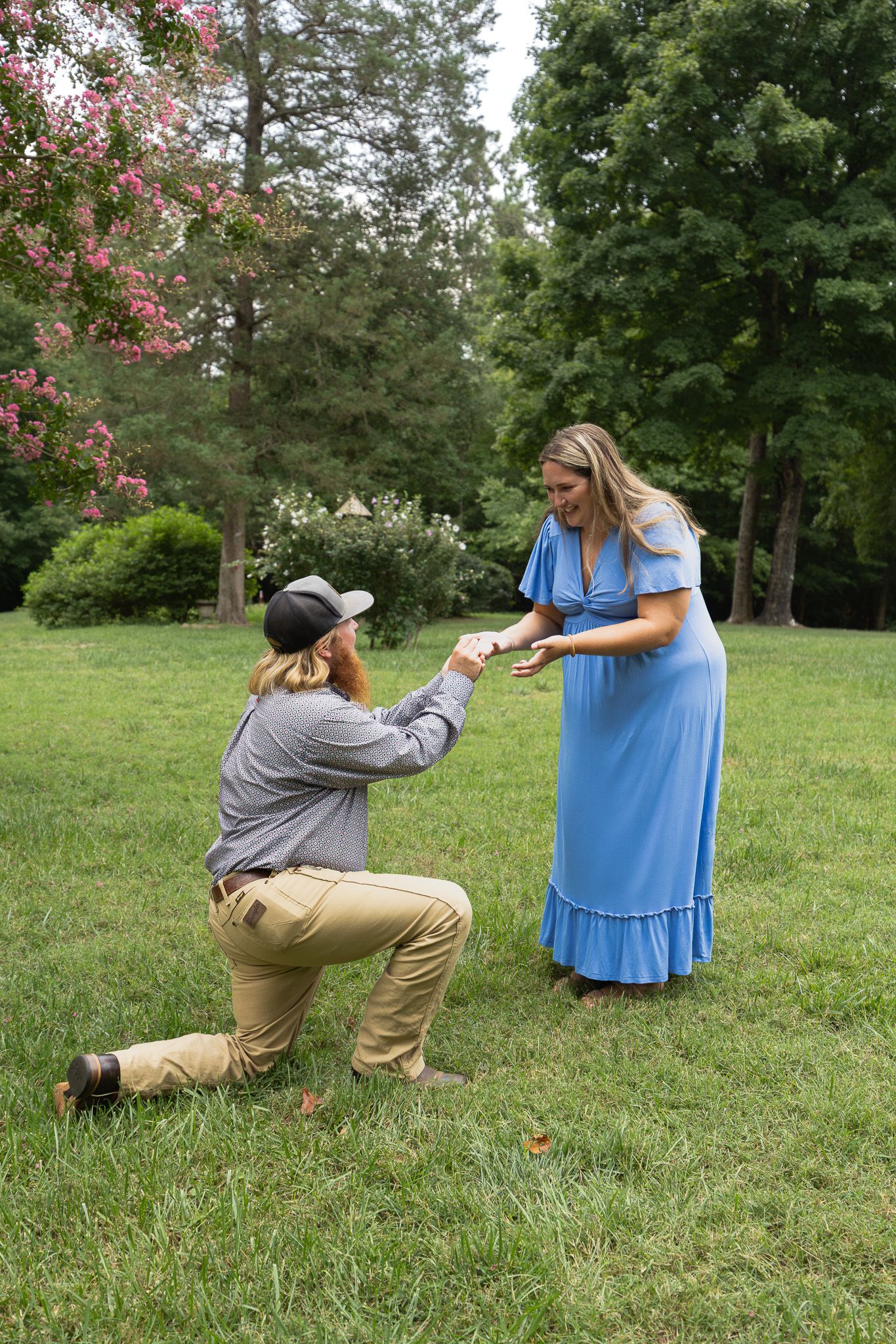Man kneeling on one knee proposing to woman in blue dress outdoors in lush green park setting.