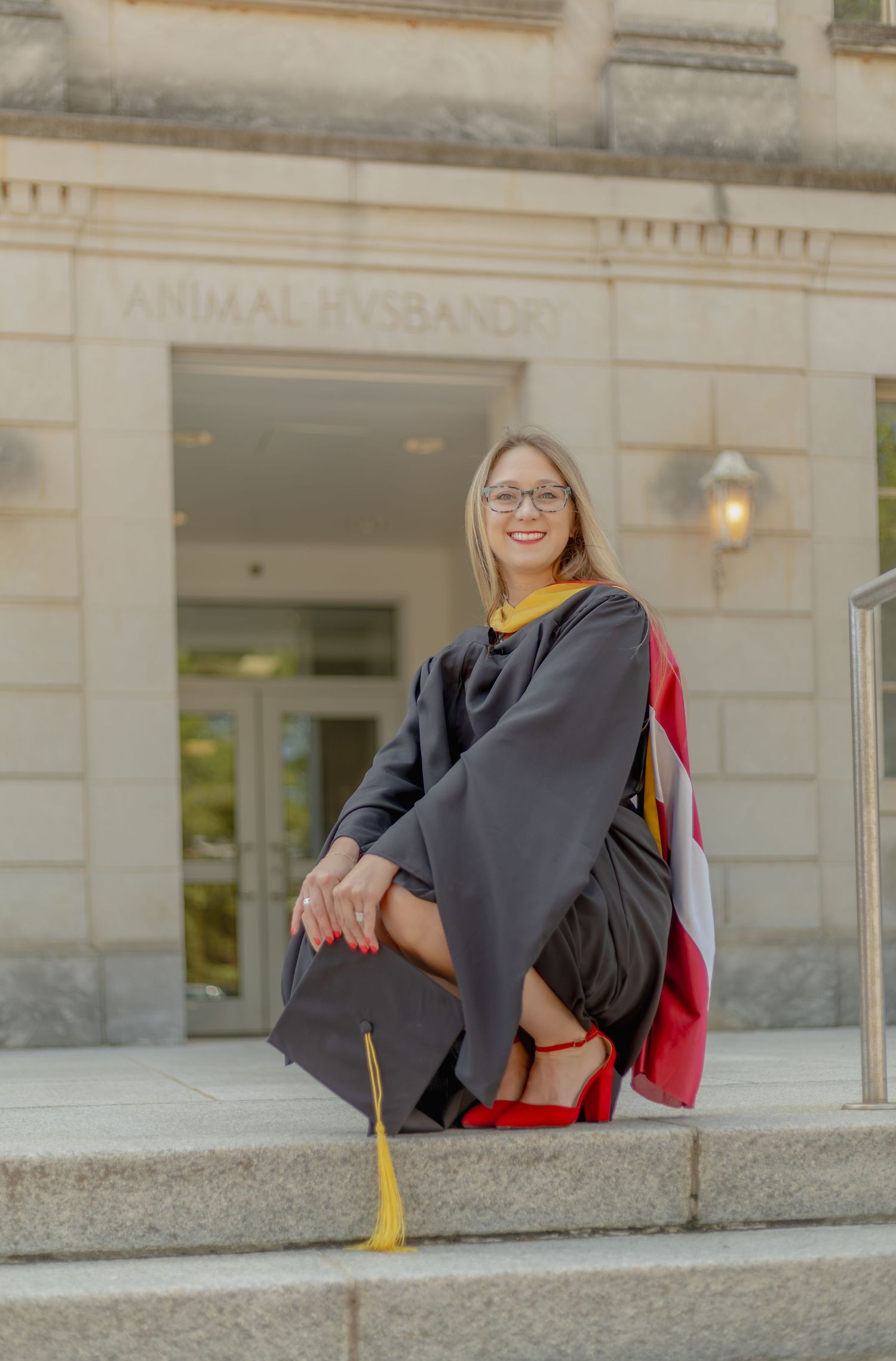 Woman in graduation gown and cap poses on steps outside Animal Husbandry building, smiling proudly.