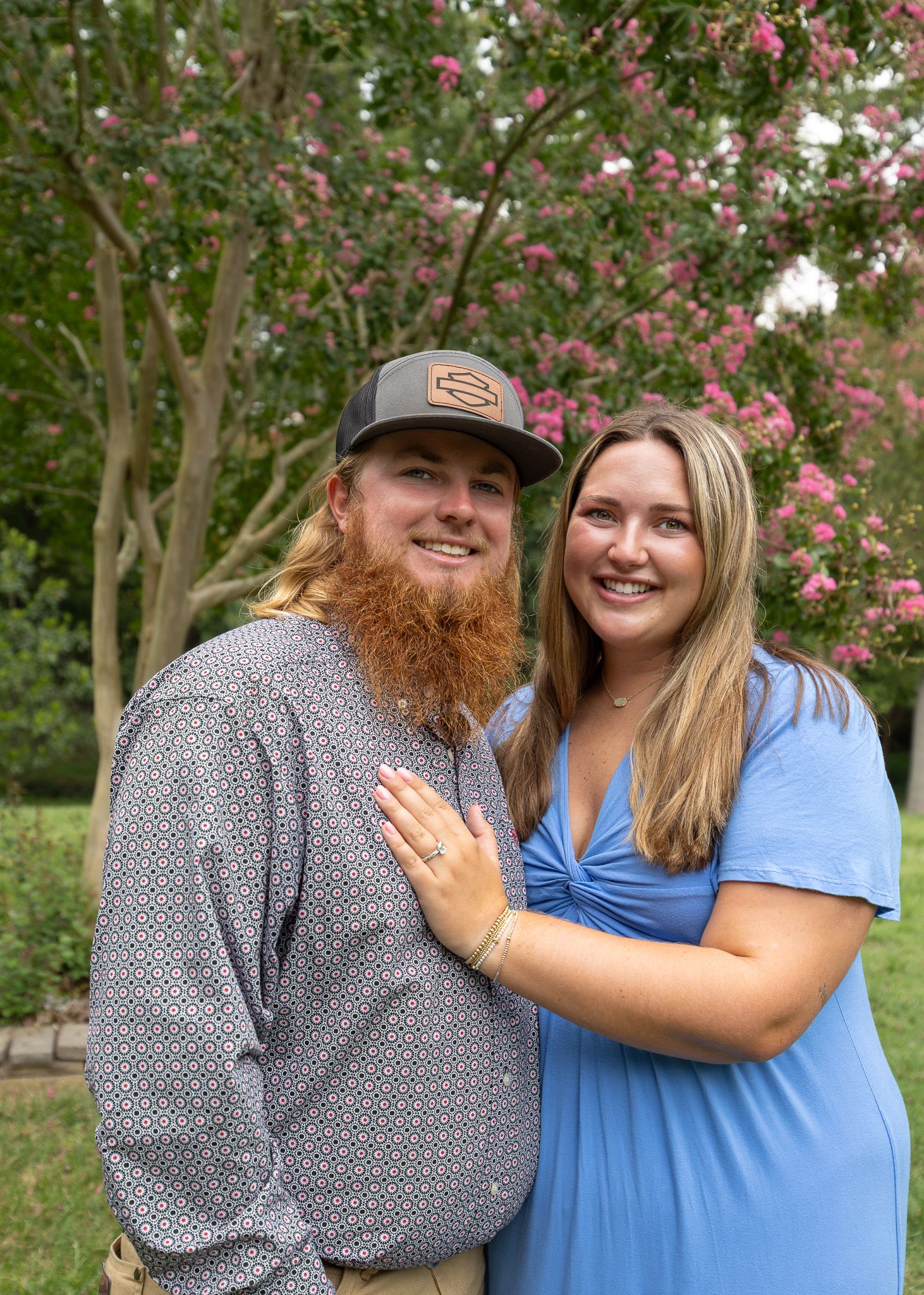 Smiling couple posing outdoors near pink flowering tree, man with red beard in cap, woman in blue dress.