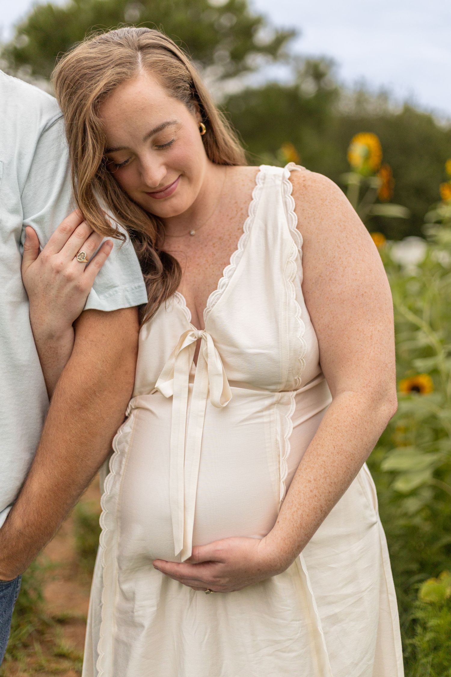 Pregnant woman in white dress tenderly holds her baby bump beside partner in sunflower field maternity photo.
