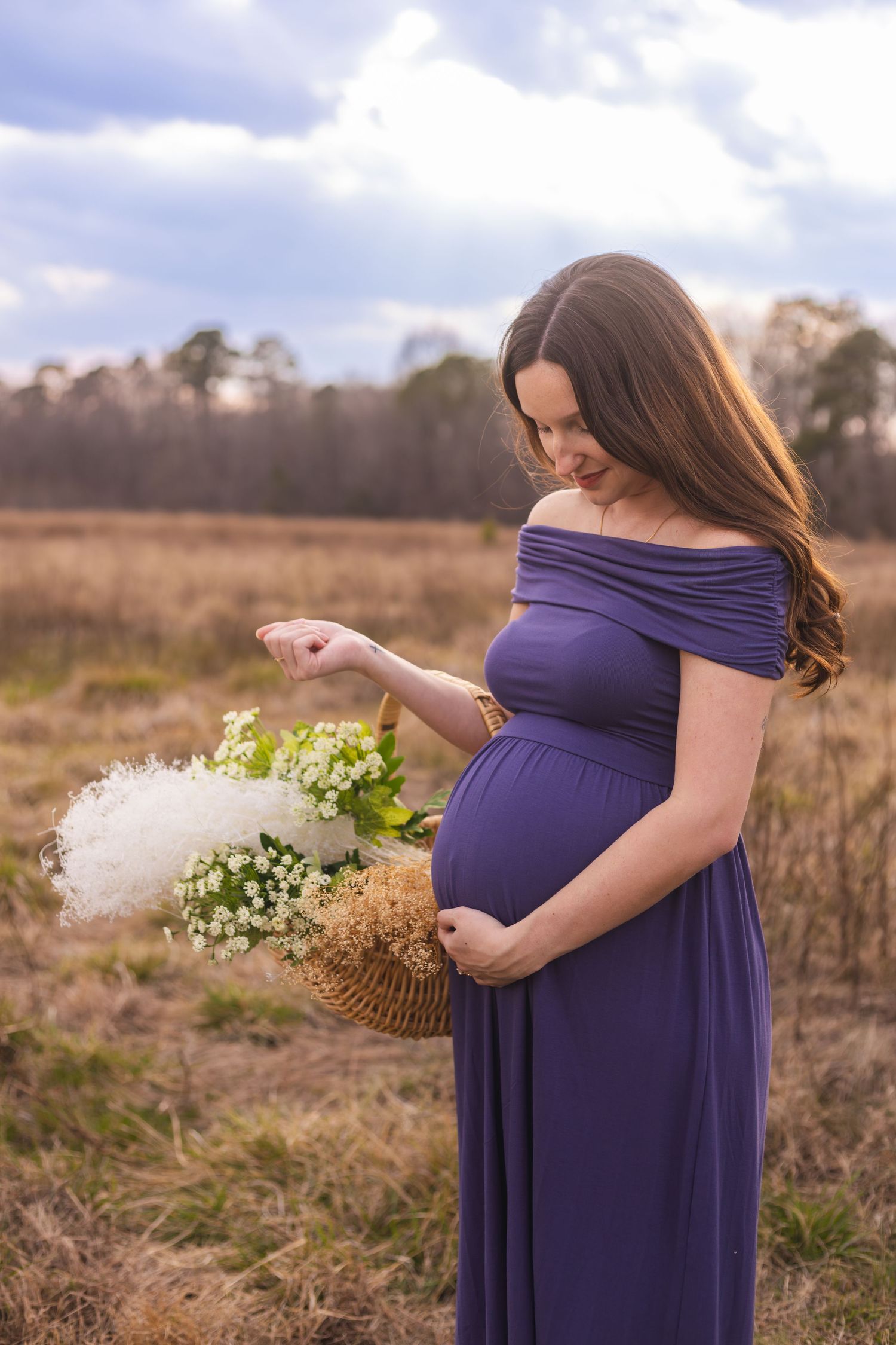 Woman in purple dress holding flowers during outdoor maternity photoshoot in a rustic field.