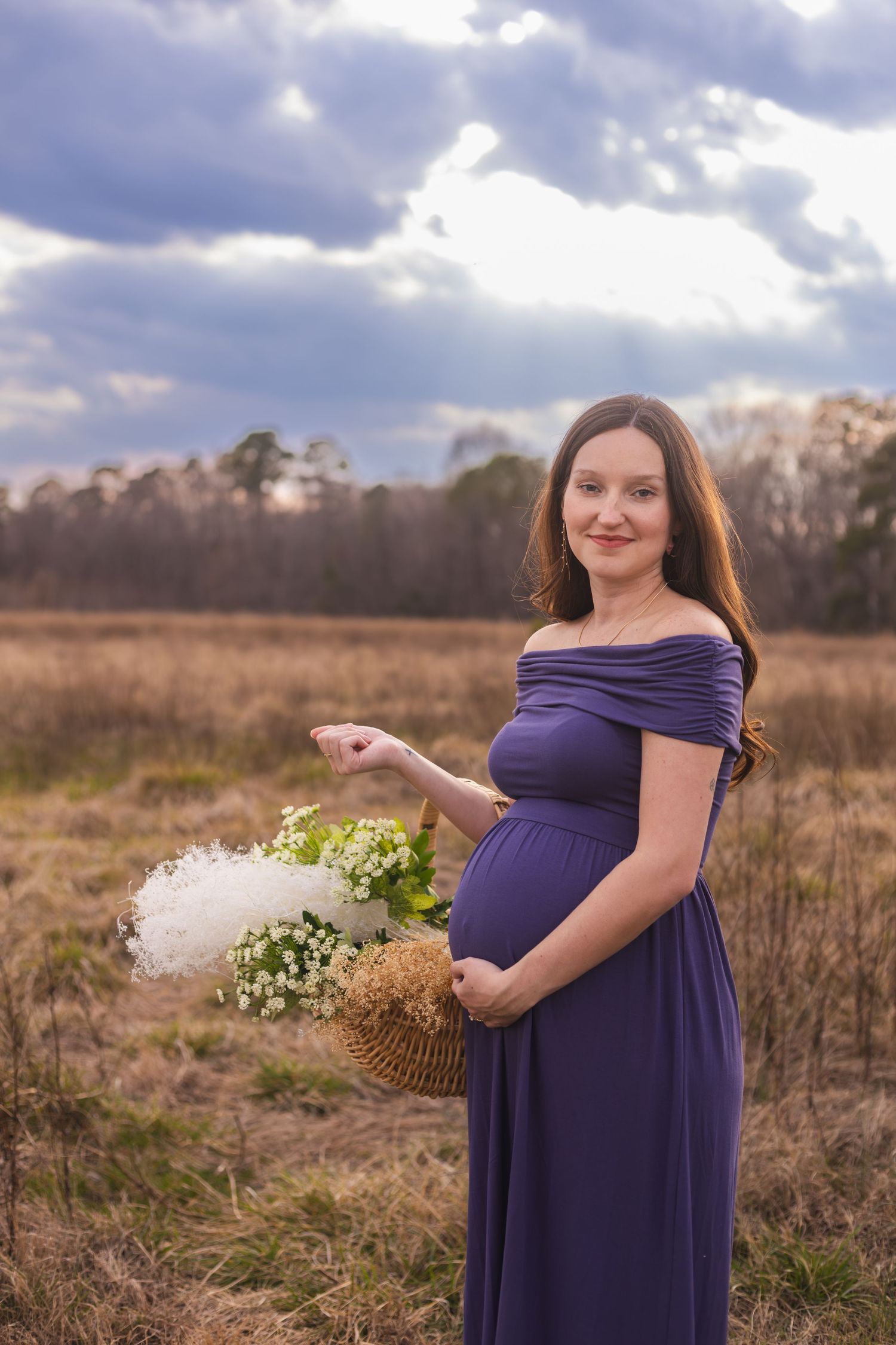 Expectant mother in purple dress poses with flowers in a rustic field setting.