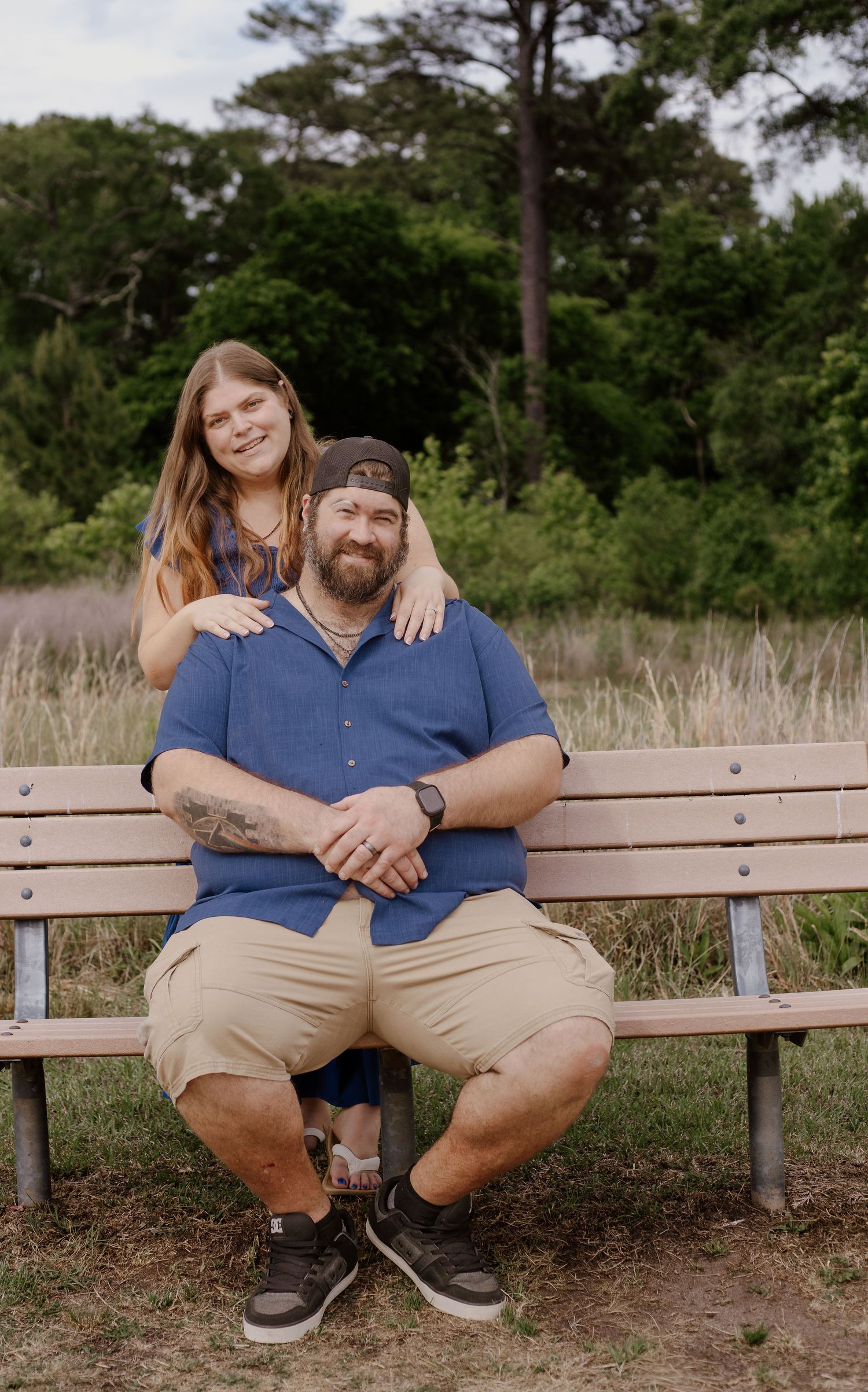 Couple posing on a wooden bench outdoors, man in blue shirt and khaki shorts, woman standing behind him smiling.