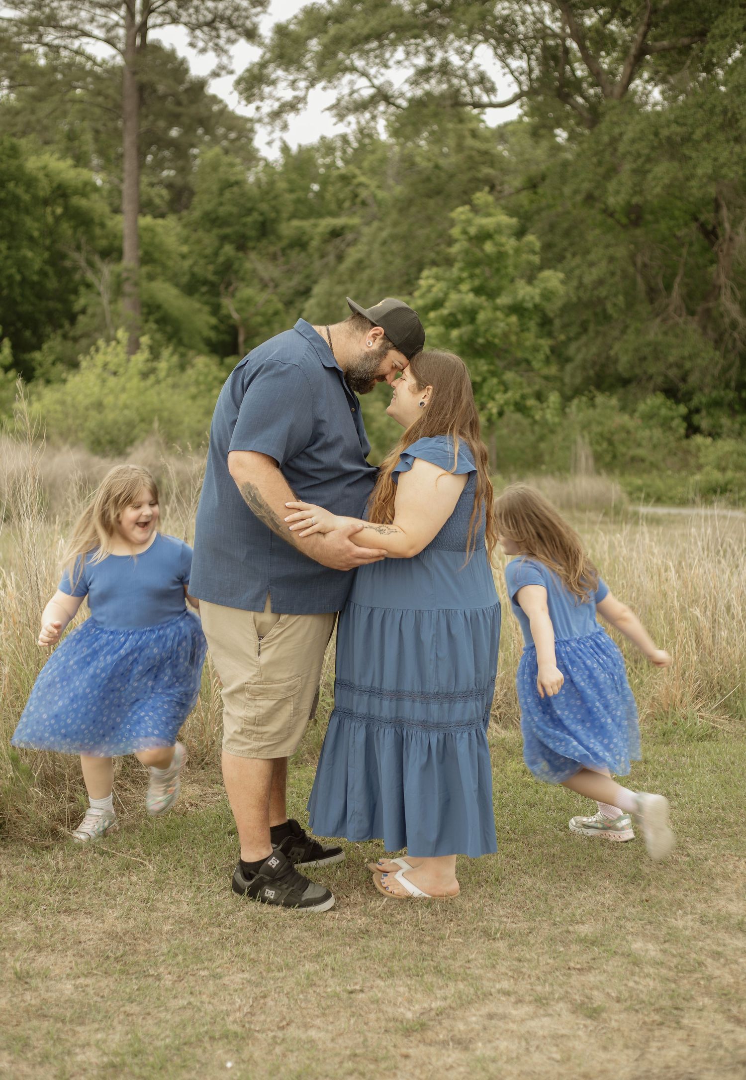 A couple shares a kiss outdoors while two young girls in matching blue dresses play around them during a family photo session.