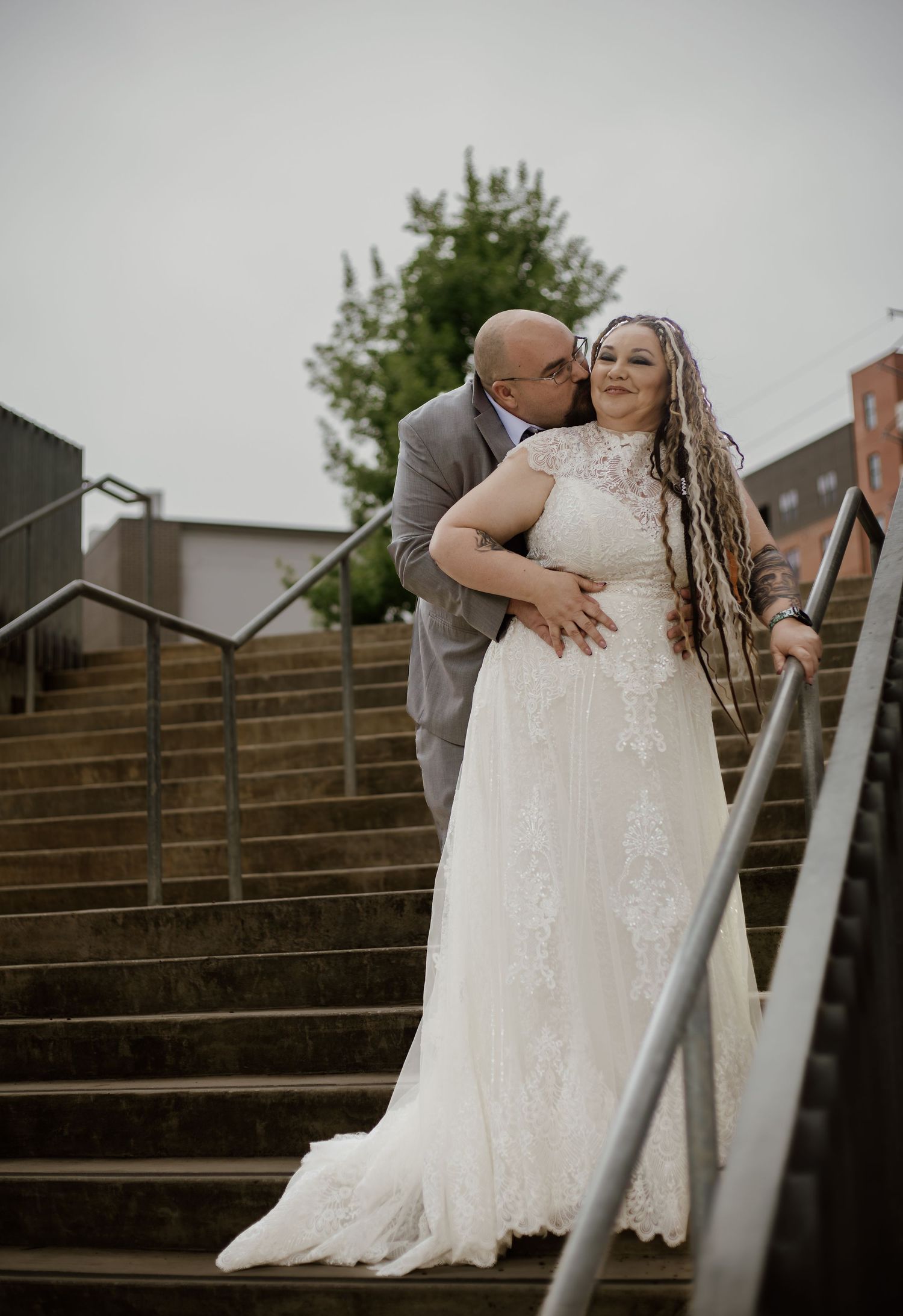 A groom kisses his bride on the cheek as they pose on outdoor steps, she wearing a lace wedding gown with long braids.