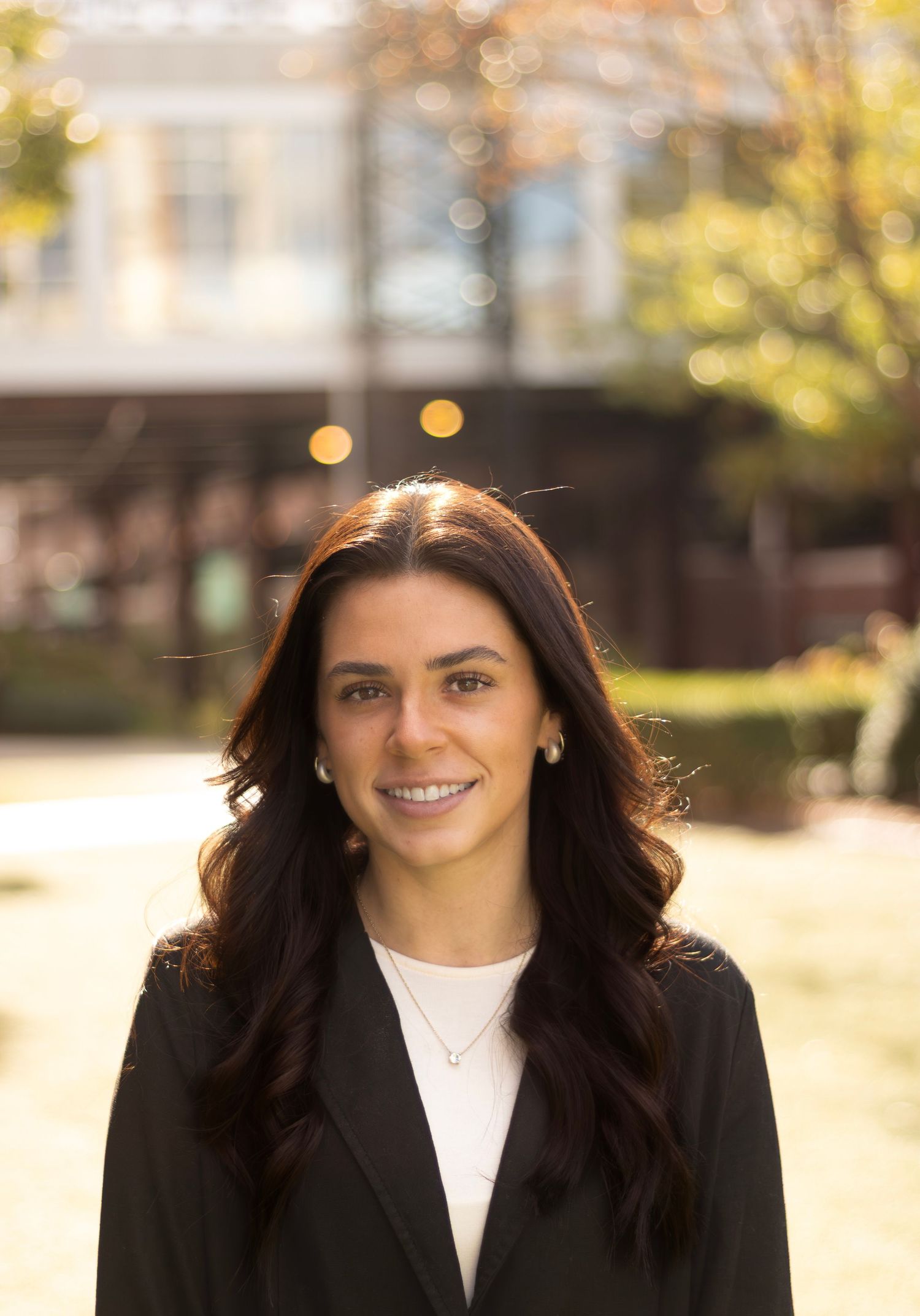 Professional headshot of someone in a black blazer and white top against an autumn background.