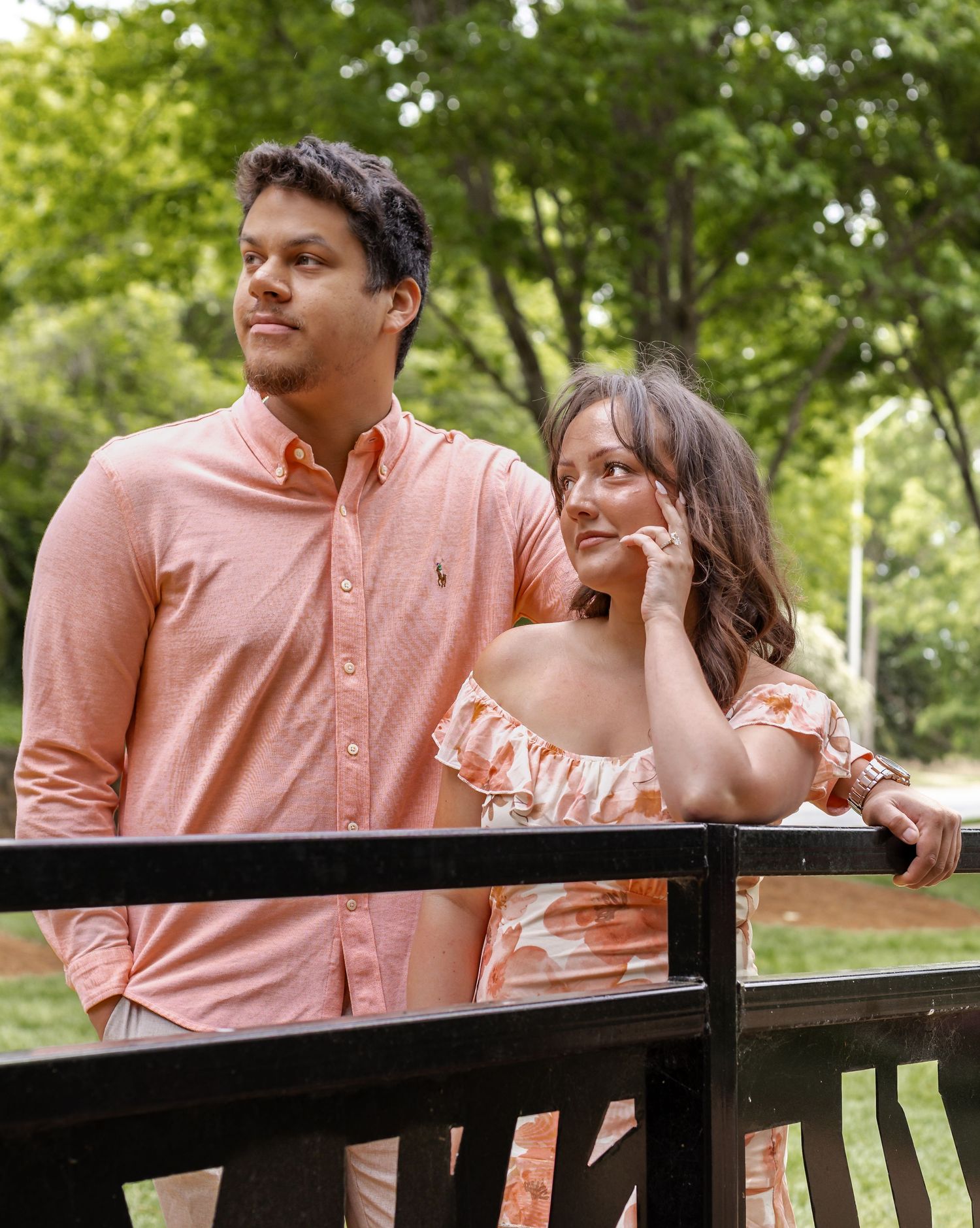 A couple in matching coral outfits poses on a wooden bridge surrounded by lush green trees.