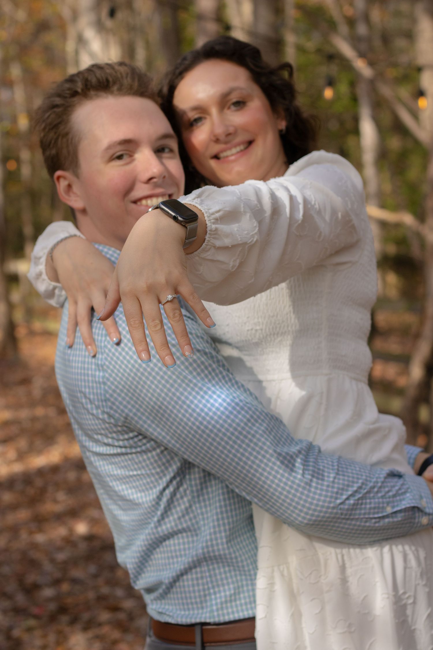 A couple embraces outdoors, with one wearing a white dress and the other in a blue checkered shirt.