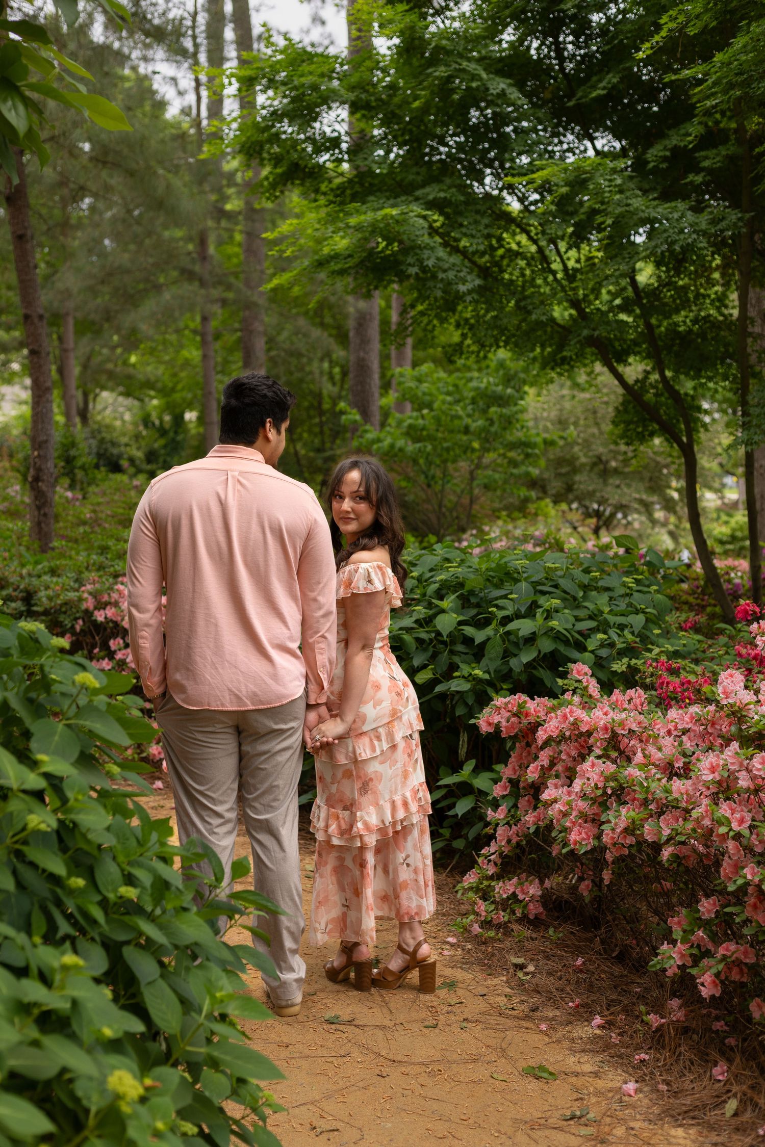 Couple holding hands walking on a garden path surrounded by lush greenery and pink flowering bushes.