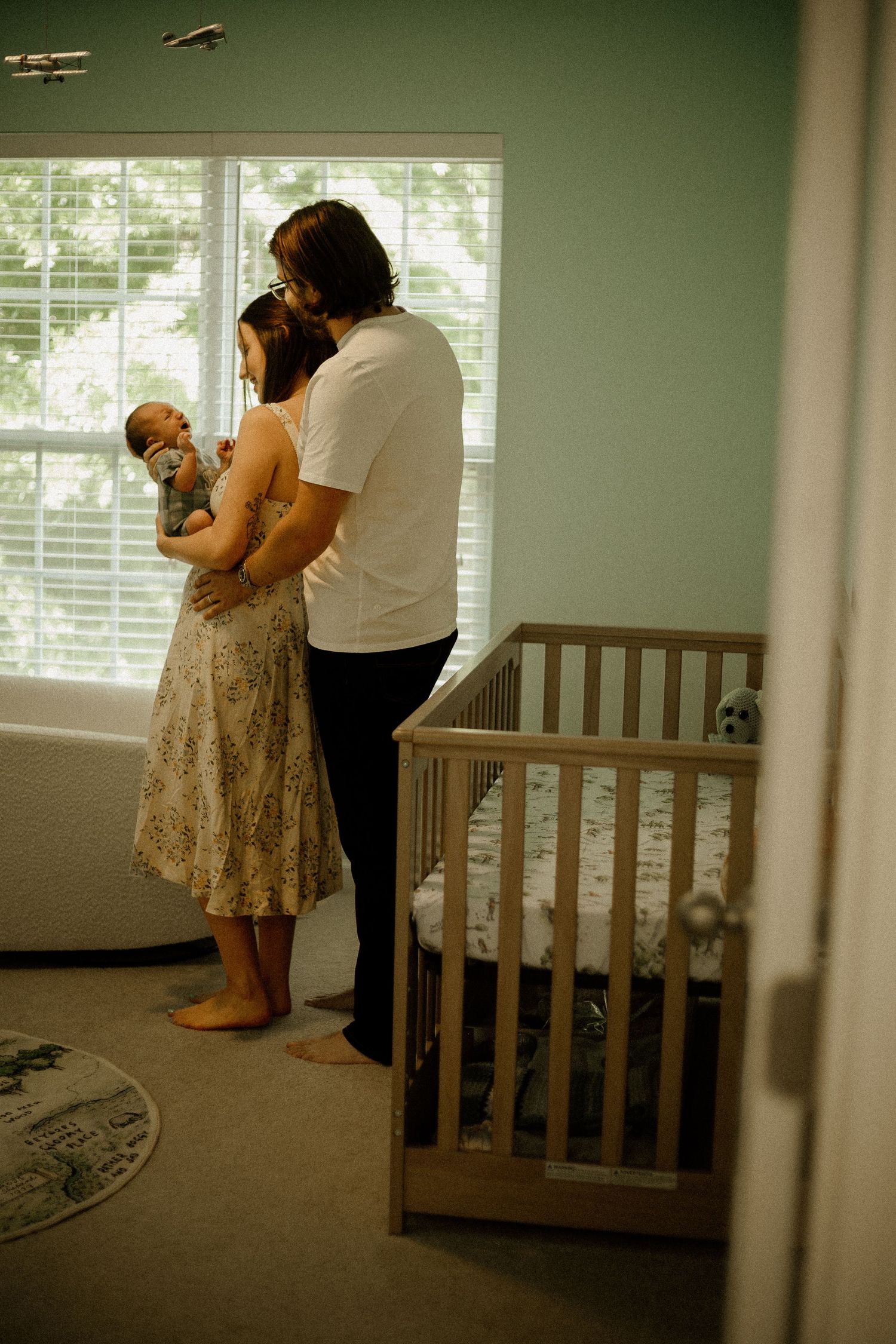 A couple stands by a nursery window holding their newborn baby beside a wooden crib in a soft green room.