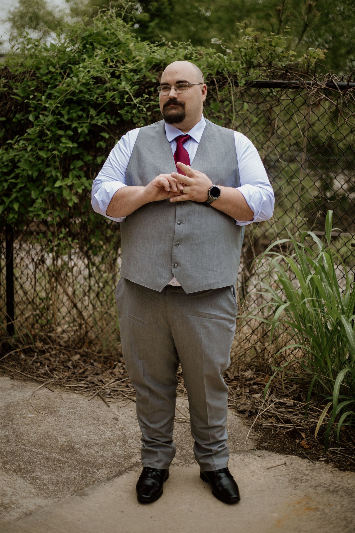 Bald man with glasses wearing a gray vest, burgundy tie, and dress pants stands outdoors near green foliage.