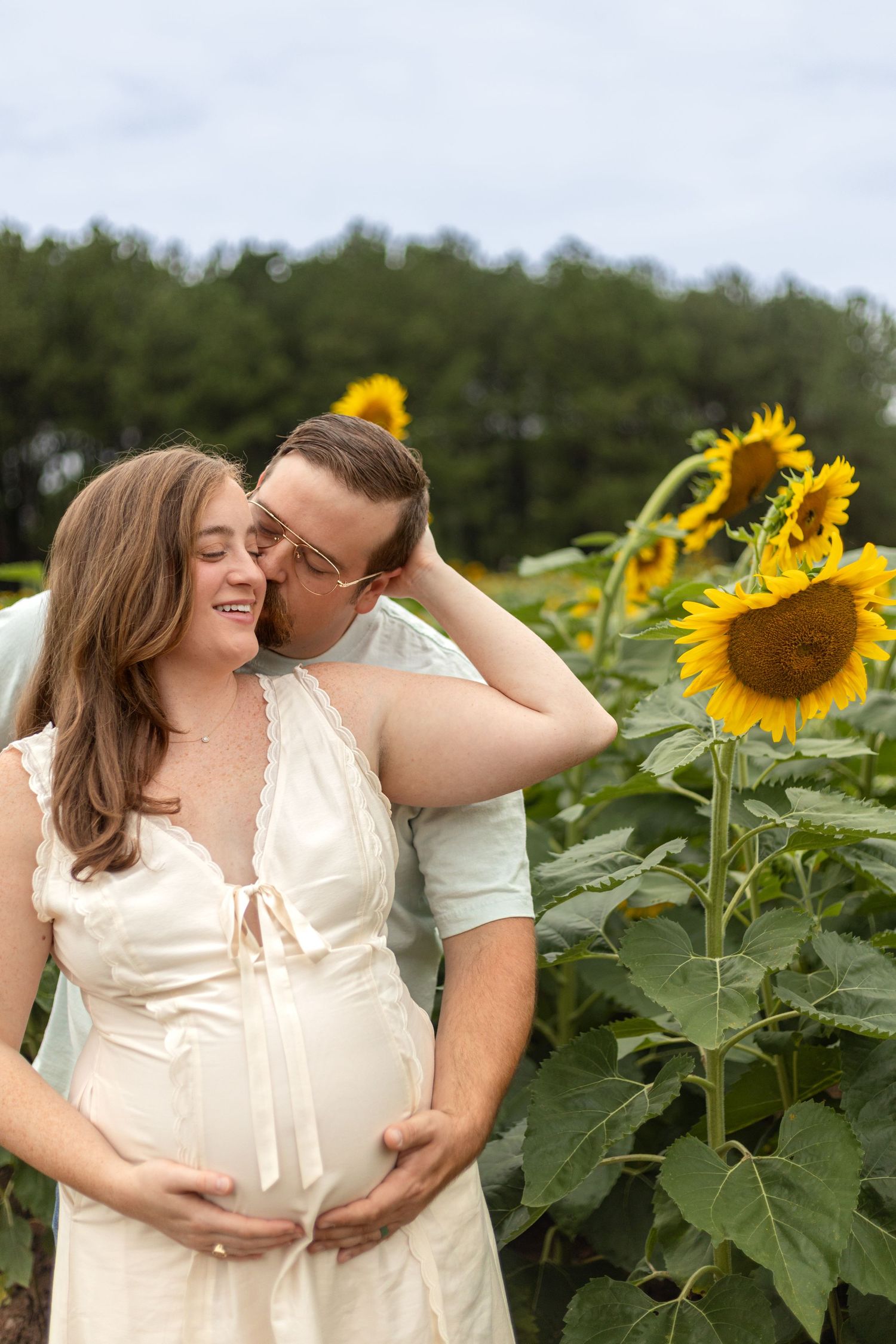 Pregnant couple embracing in a sunflower field, woman in white dress smiling as man kisses her cheek.
