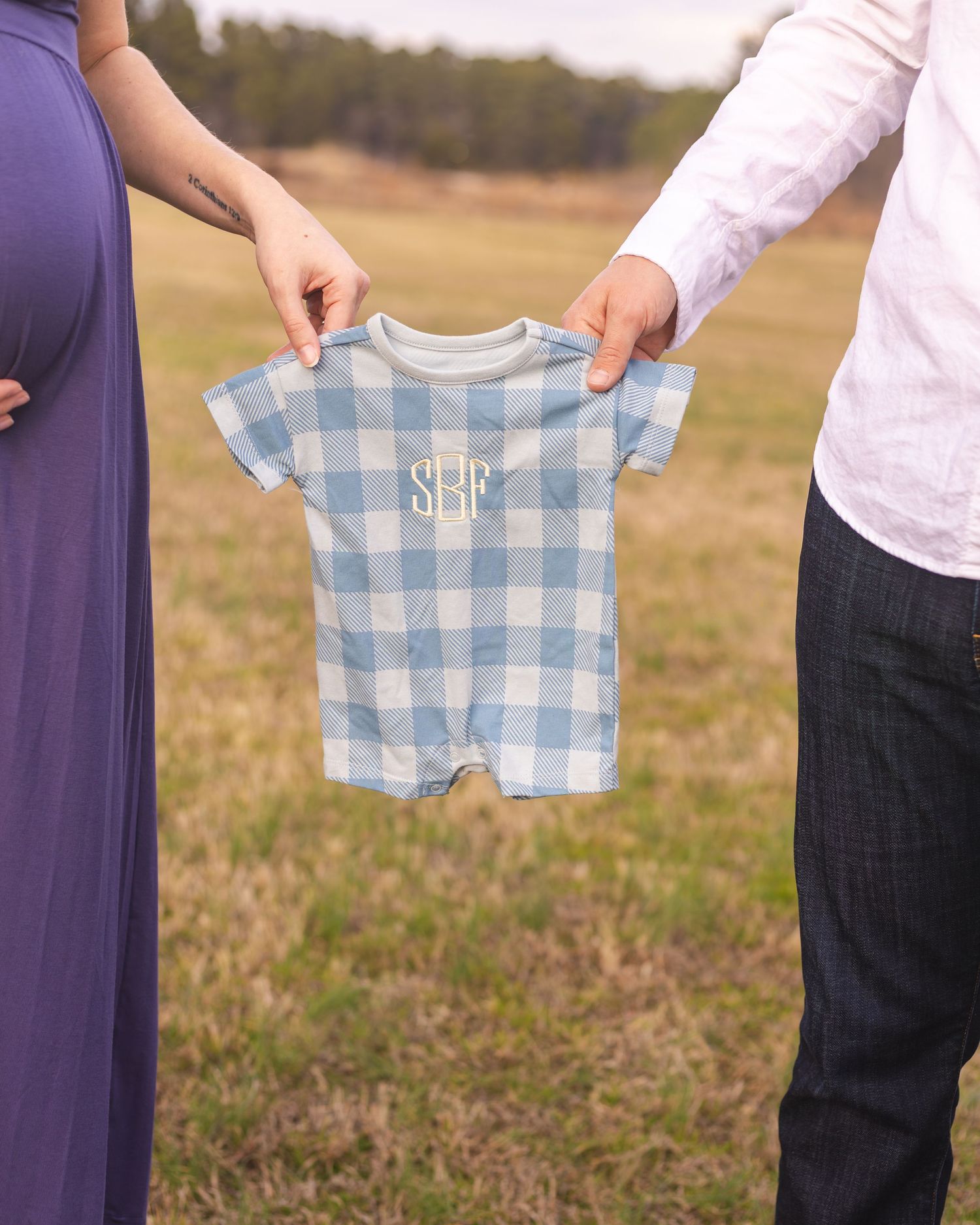 Couple holding up tiny blue gingham baby clothes in an outdoor field setting.