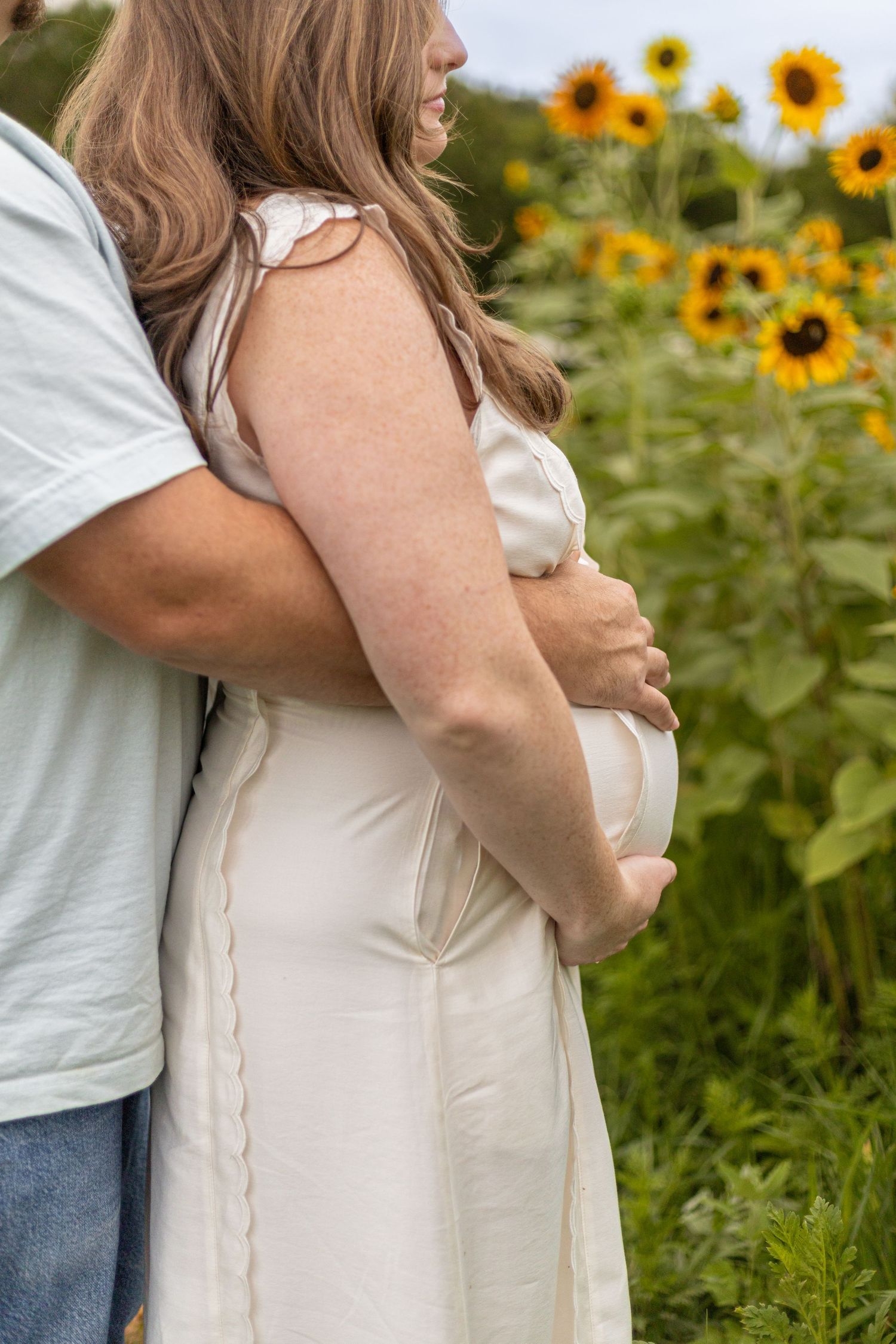 Pregnant woman in white dress embraced by partner in sunflower field during maternity photo session.