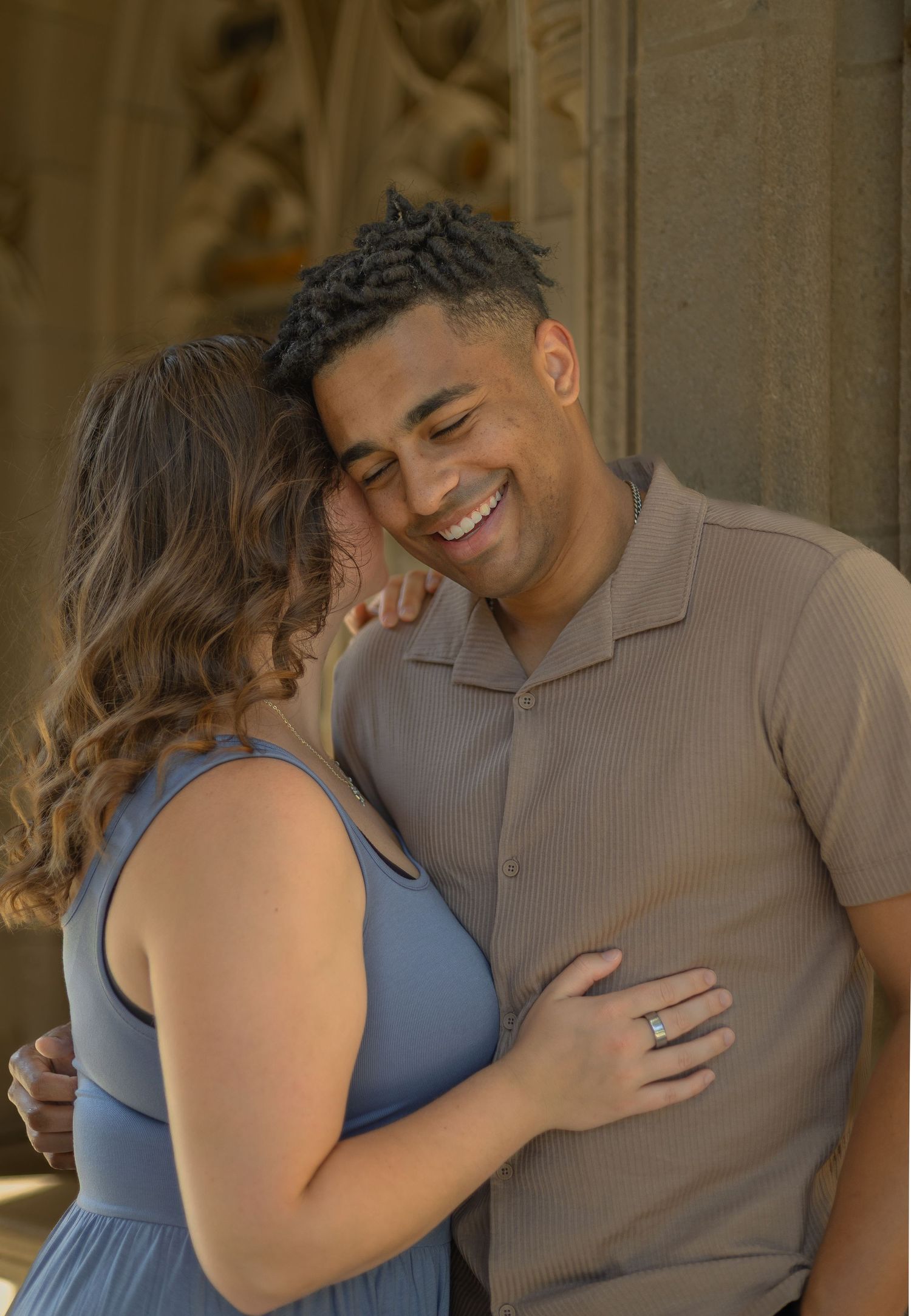 A smiling couple embraces warmly, the woman wearing a blue dress and an engagement ring, against ornate stone columns.