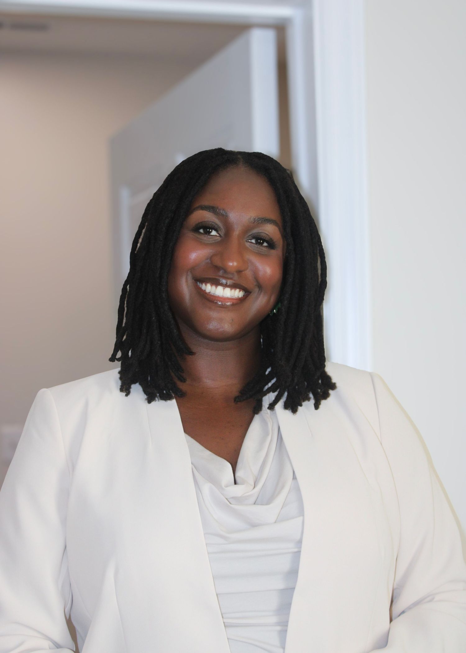 Professional wearing a white blazer and cowl neck top smiles warmly against a light background.
