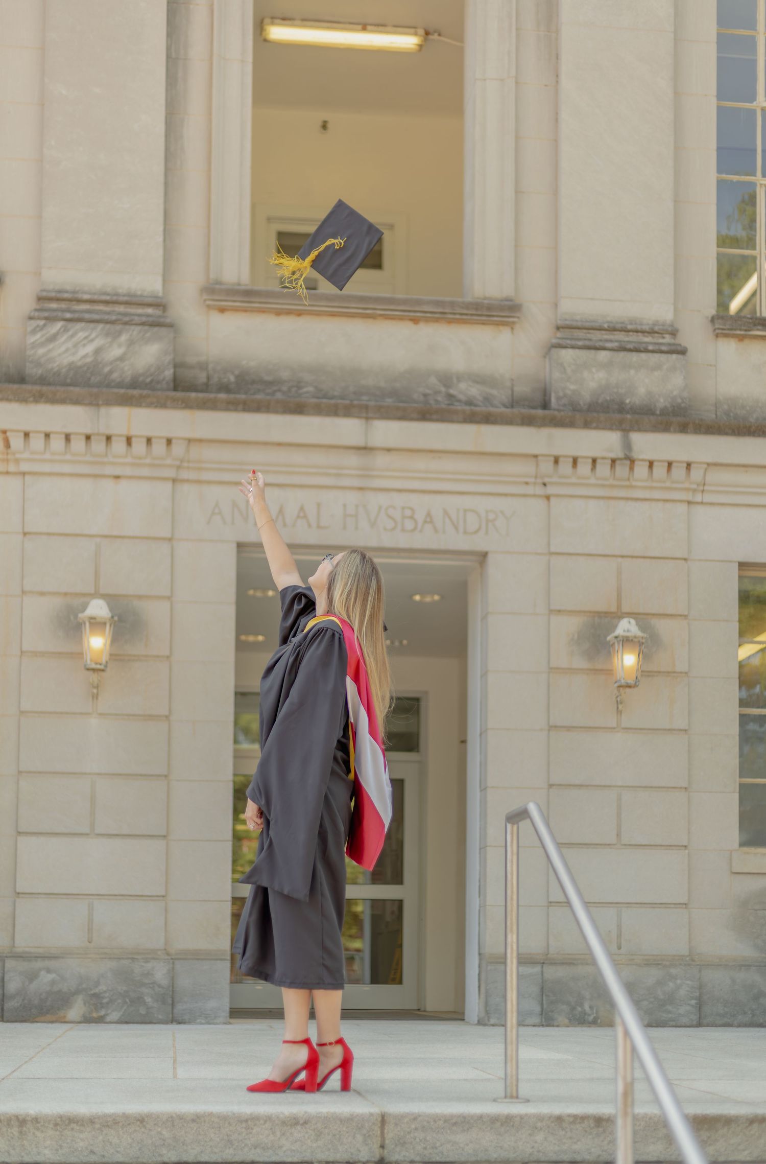 Graduate in black gown and red heels tosses cap outside Animal Husbandry building during graduation celebration.