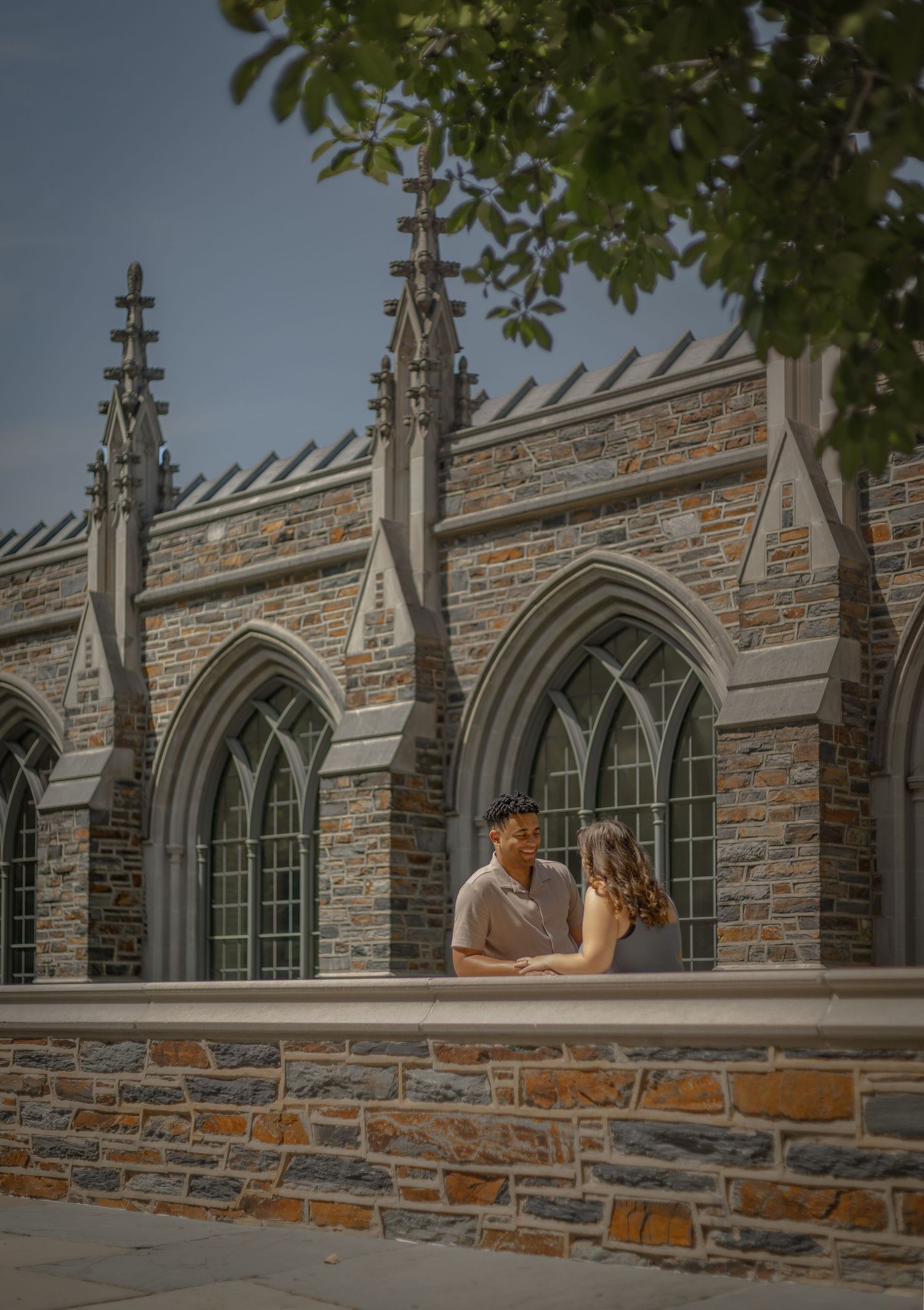 Couple embracing in front of a Gothic stone building with arched windows and decorative spires on a sunny day.
