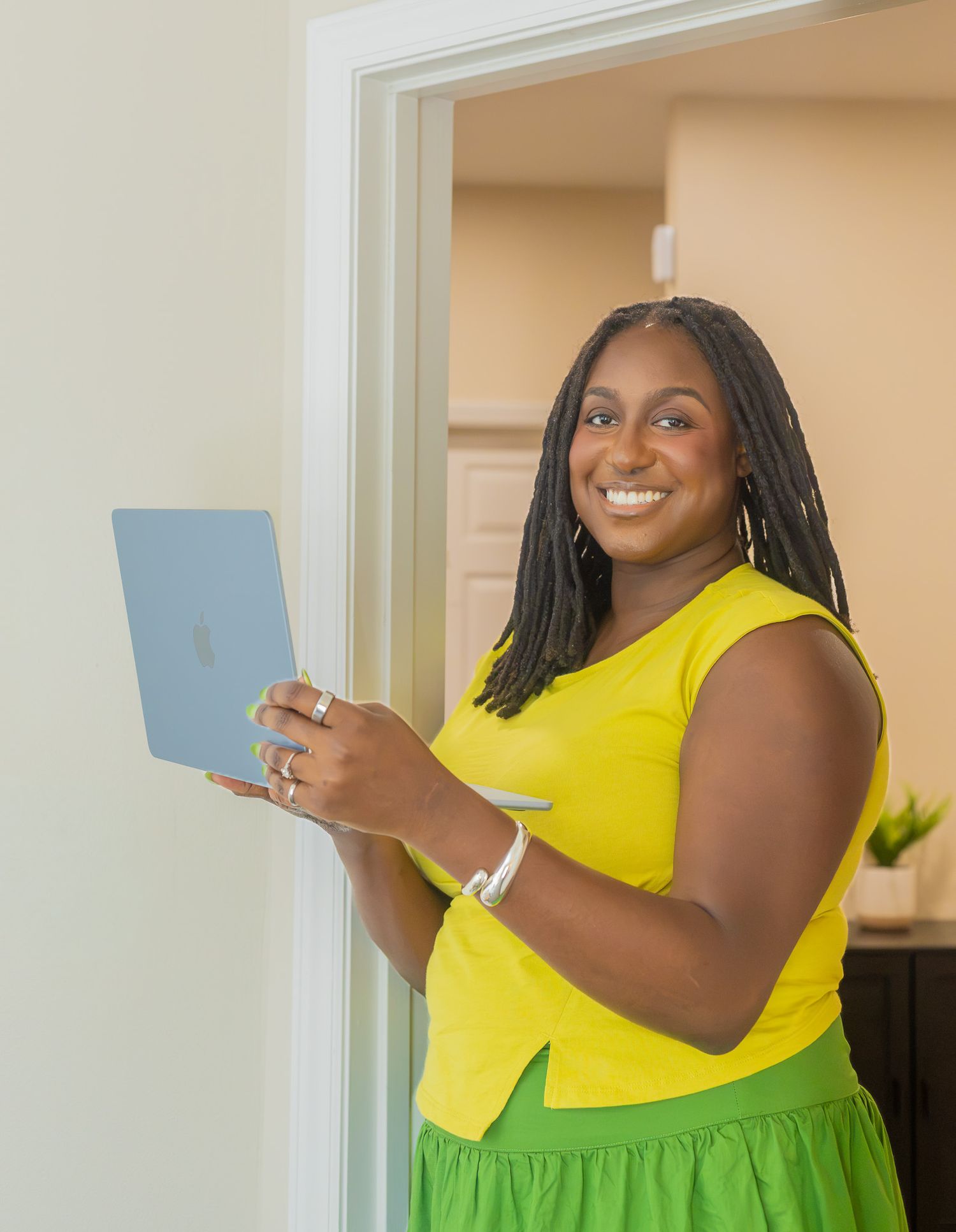 Smiling woman in yellow top and green skirt holding a blue laptop, standing in a bright doorway.
