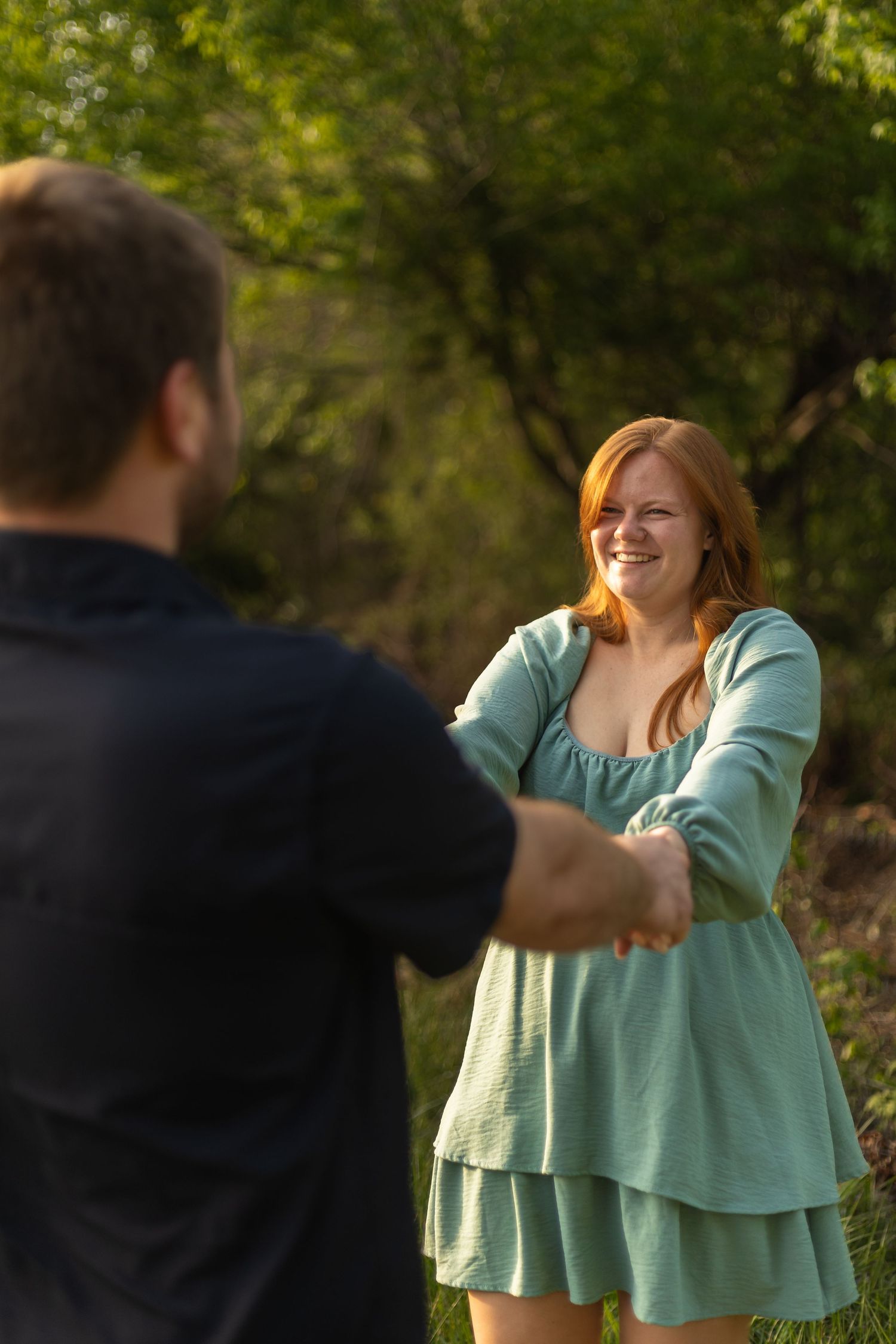A smiling redhead woman in a teal dress holds hands with a man outdoors in a sunlit green forest setting.