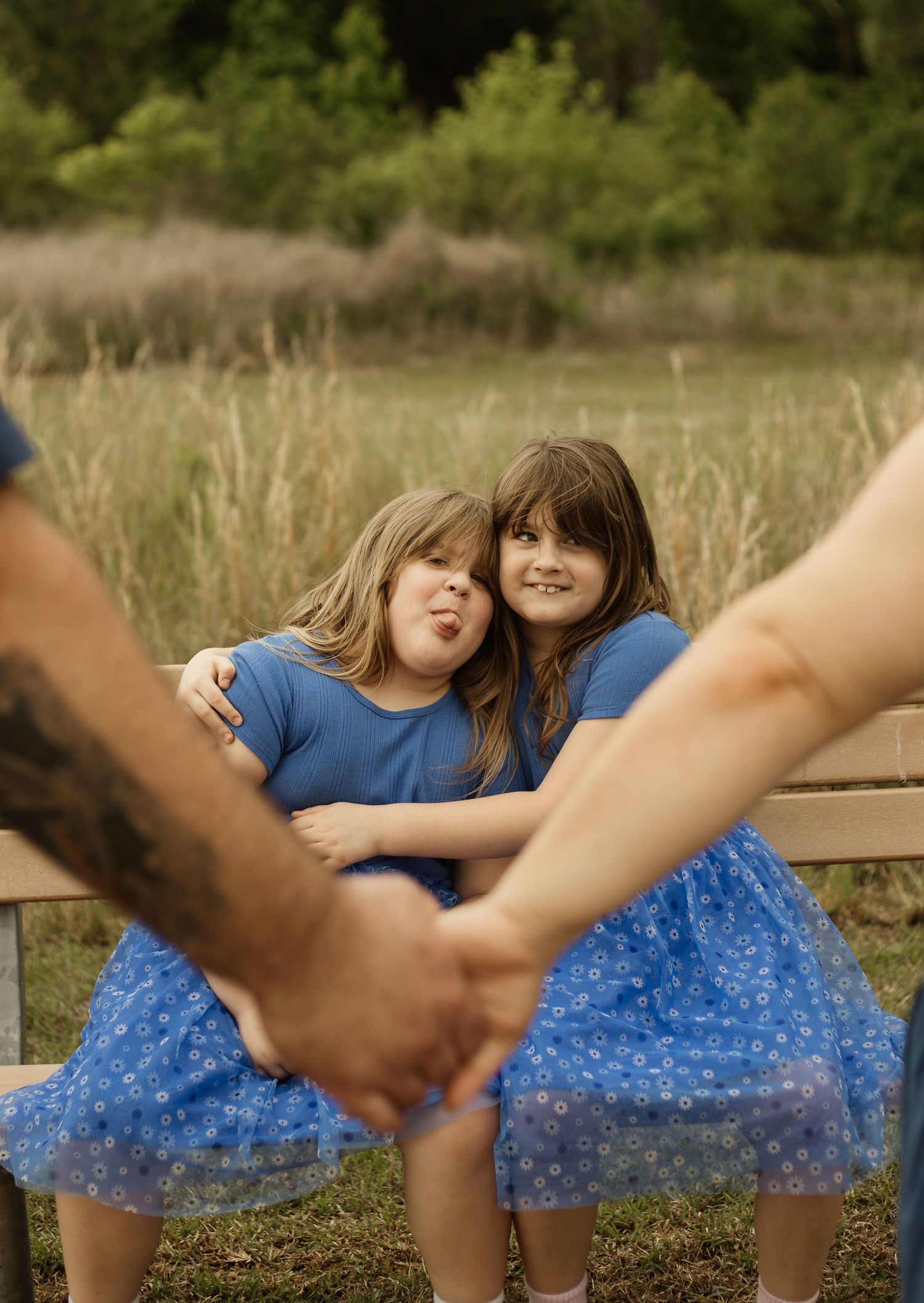 Two young girls in matching blue outfits sit on a bench outdoors, one making a silly face while adults hold hands in foreground.
