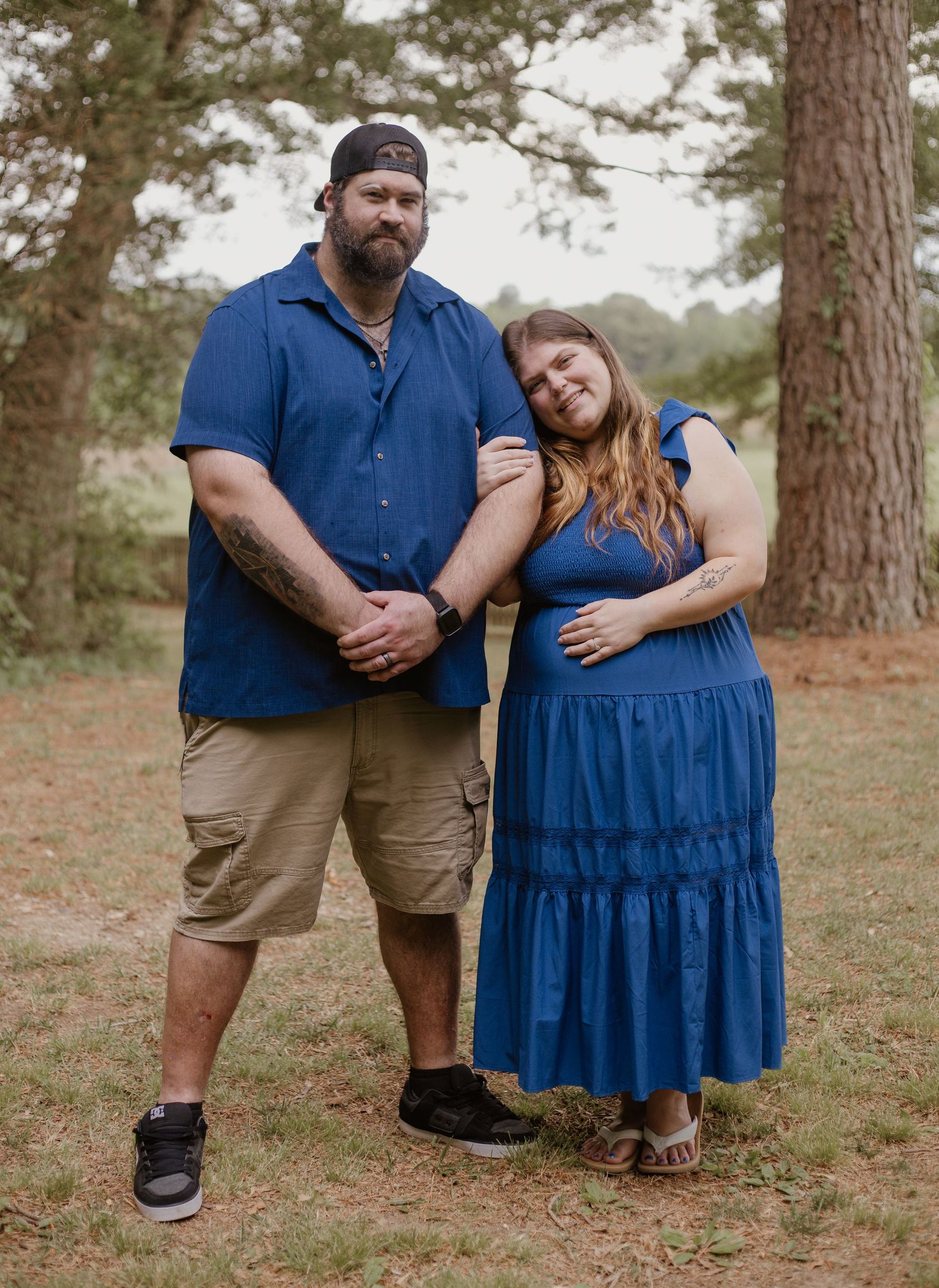 Couple in matching blue outfits posing outdoors among pine trees, woman leaning on man's shoulder.