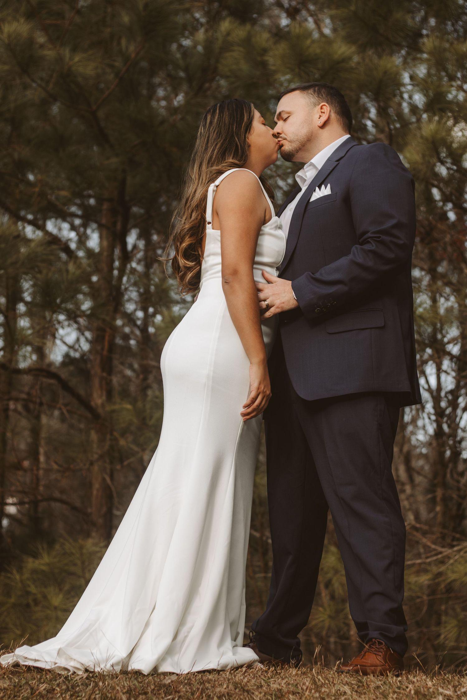 A couple in wedding attire shares an intimate moment outdoors among trees.
