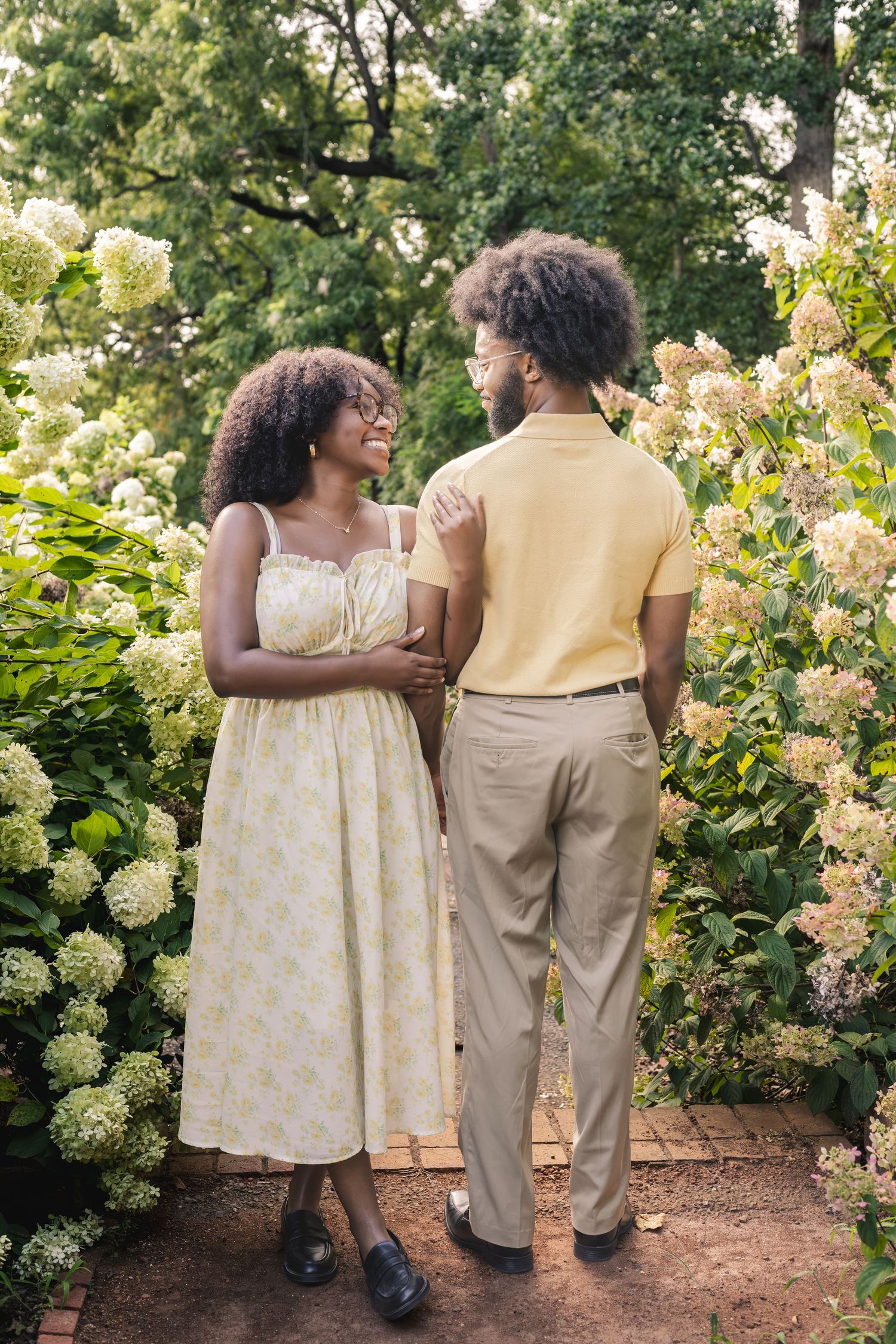 A couple walks through a lush garden with white hydrangeas, woman in floral sundress, man in yellow polo and khakis.