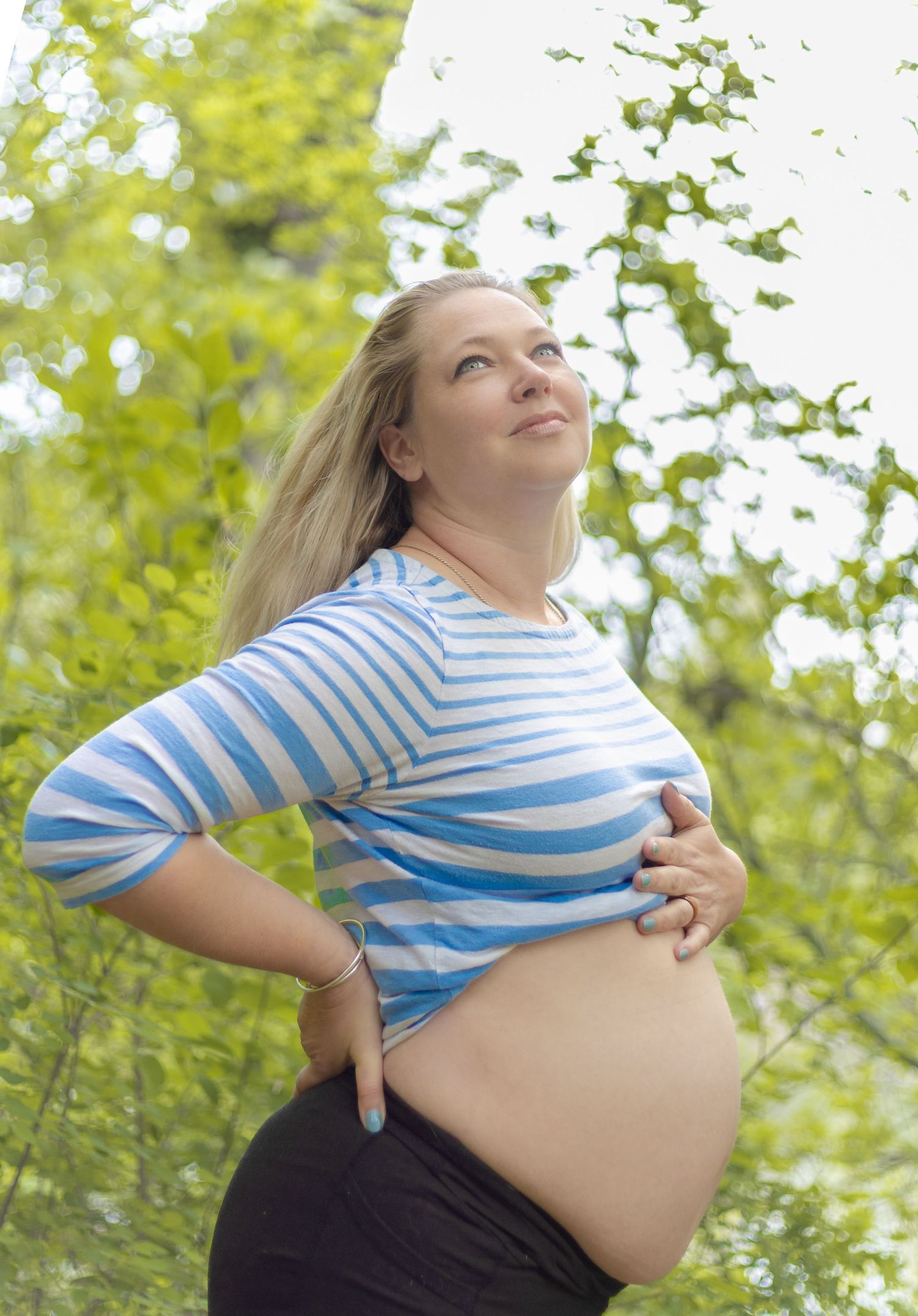 Pregnant woman in striped blue top holding her baby bump outdoors among green trees on a sunny day.
