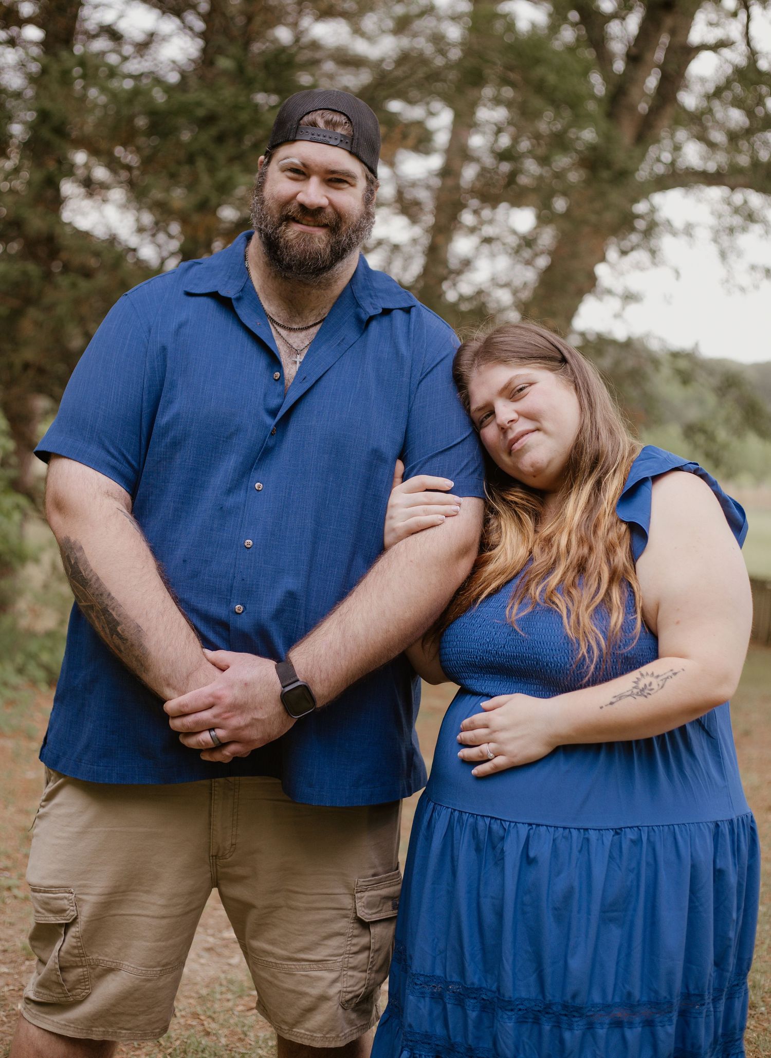 A couple in matching blue outfits poses outdoors, the woman leaning on the mans arm in a natural wooded setting.