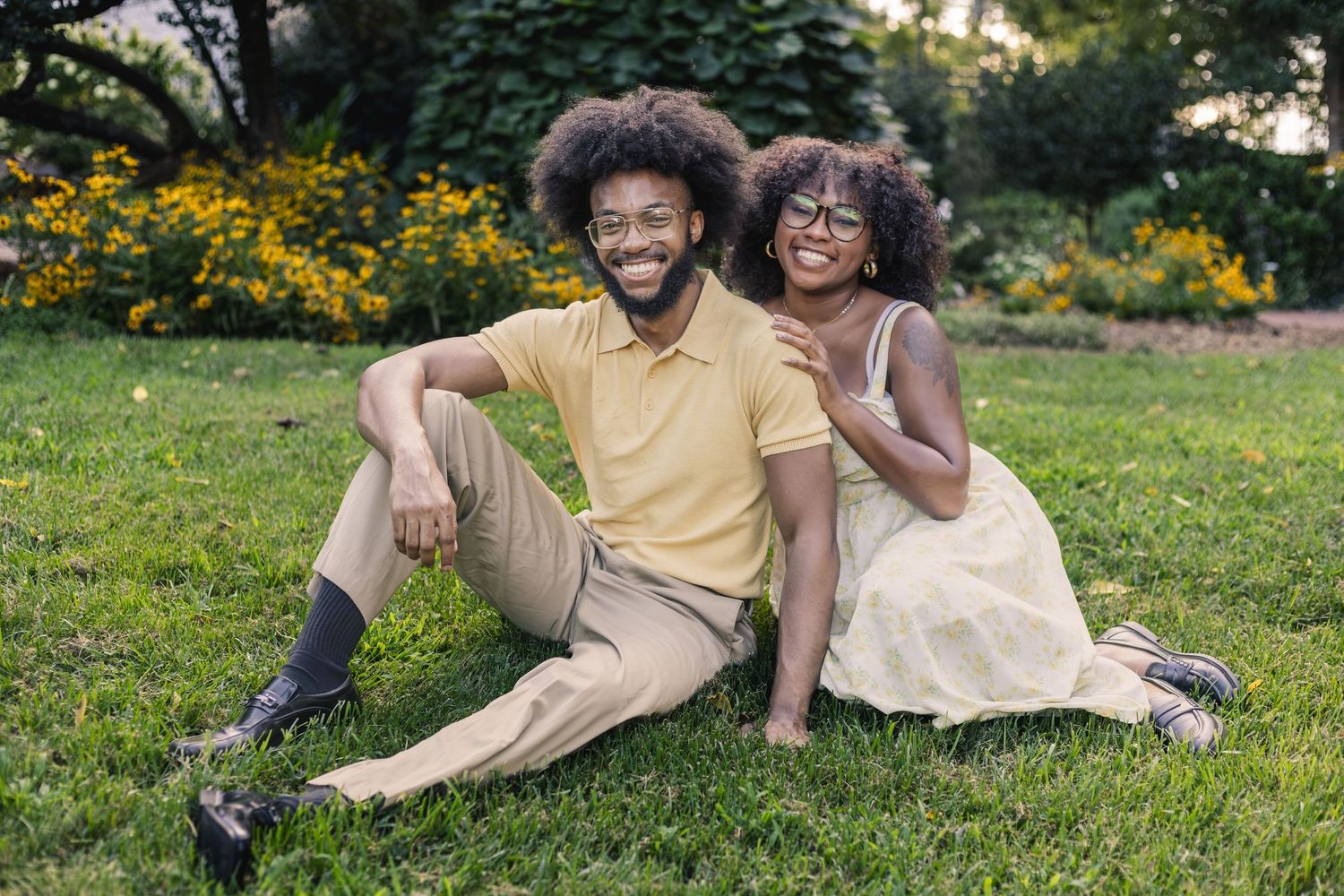 A smiling couple sits together on green grass in a park, surrounded by colorful flowers on a sunny day.