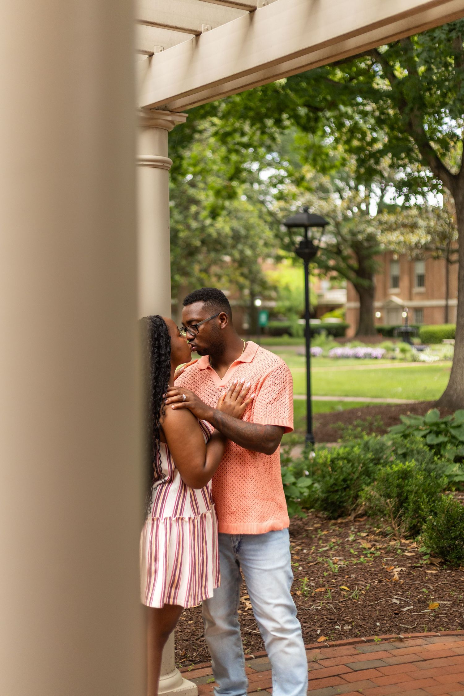 A couple shares a romantic moment by a column in a lush garden, man in pink shirt and woman in striped dress.