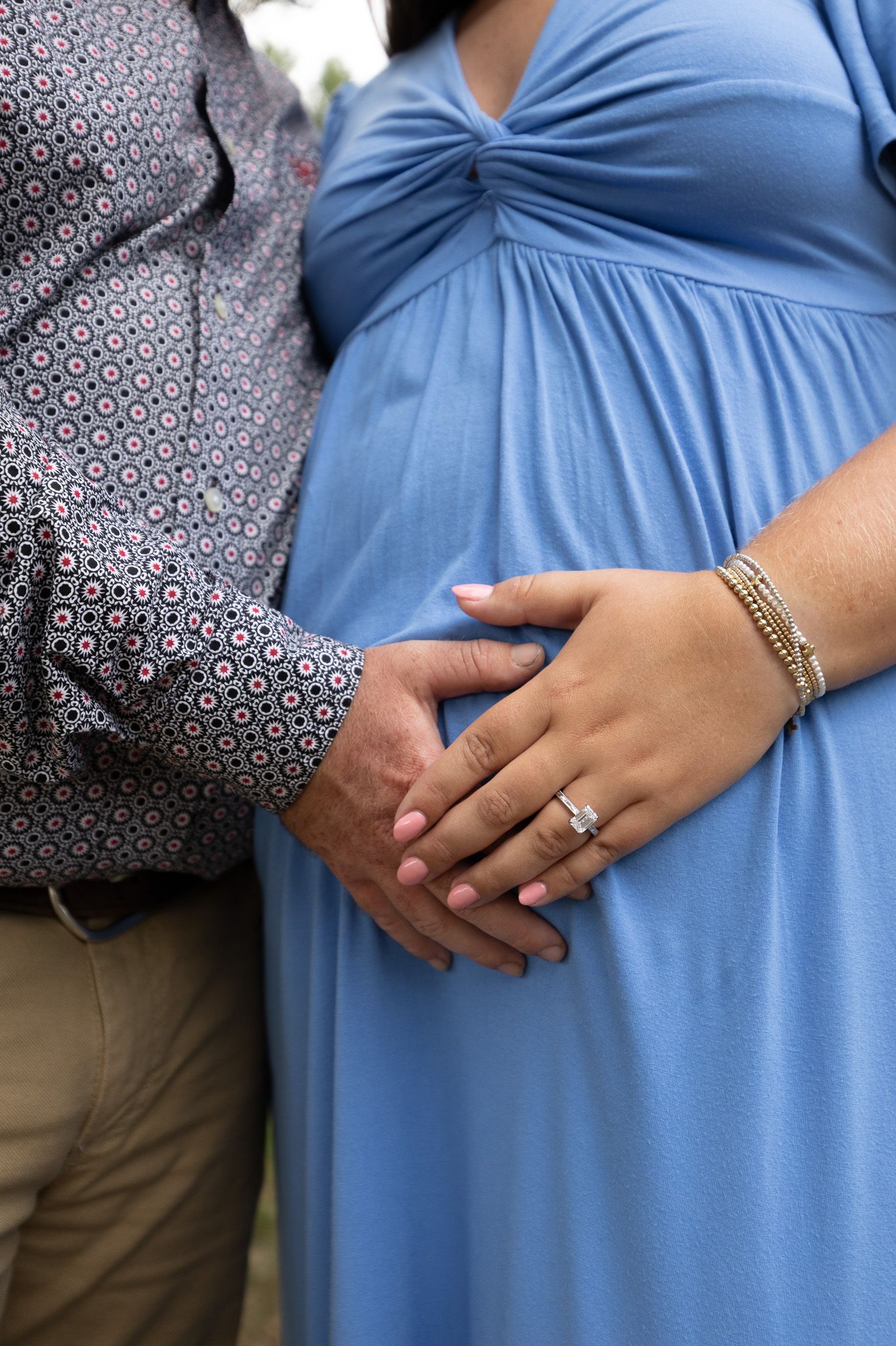 Pregnant woman in blue dress with partner's hand on baby bump, wearing diamond ring and gold bracelets.
