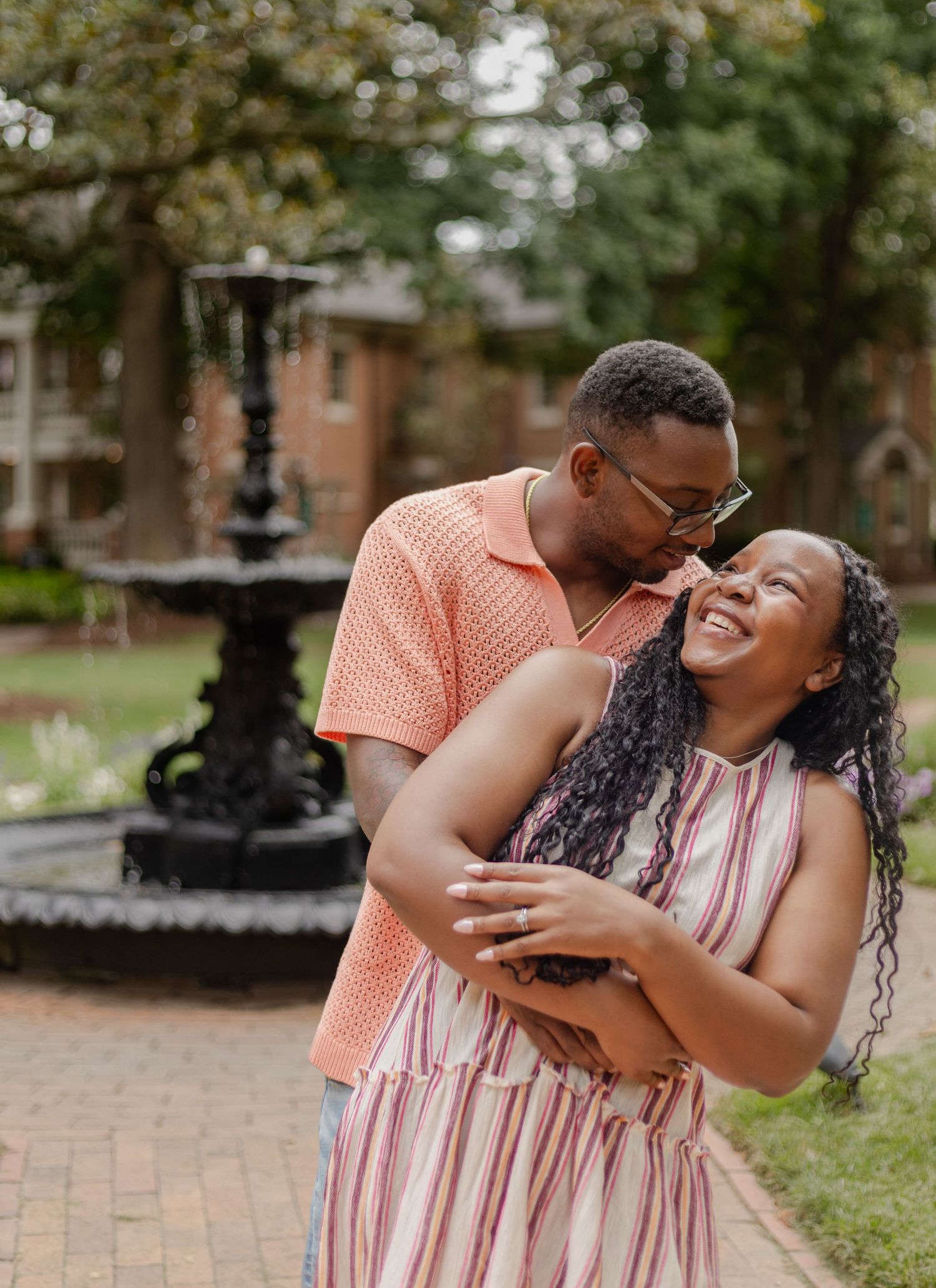 A joyful couple embraces near a vintage fountain, the man kissing her cheek as she laughs, both dressed in summer attire.