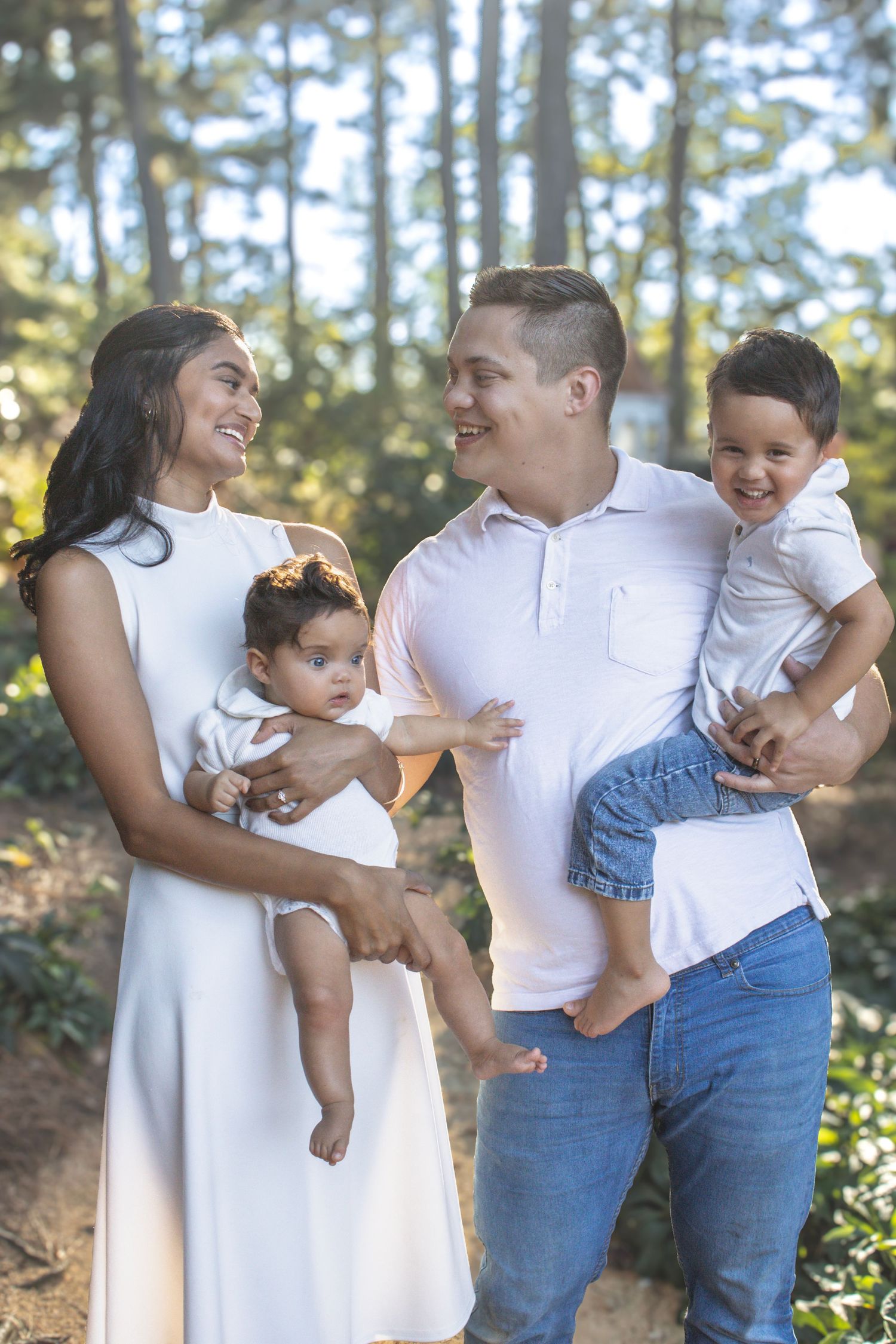 Happy family in white outfits playing and laughing together in a pine forest setting.