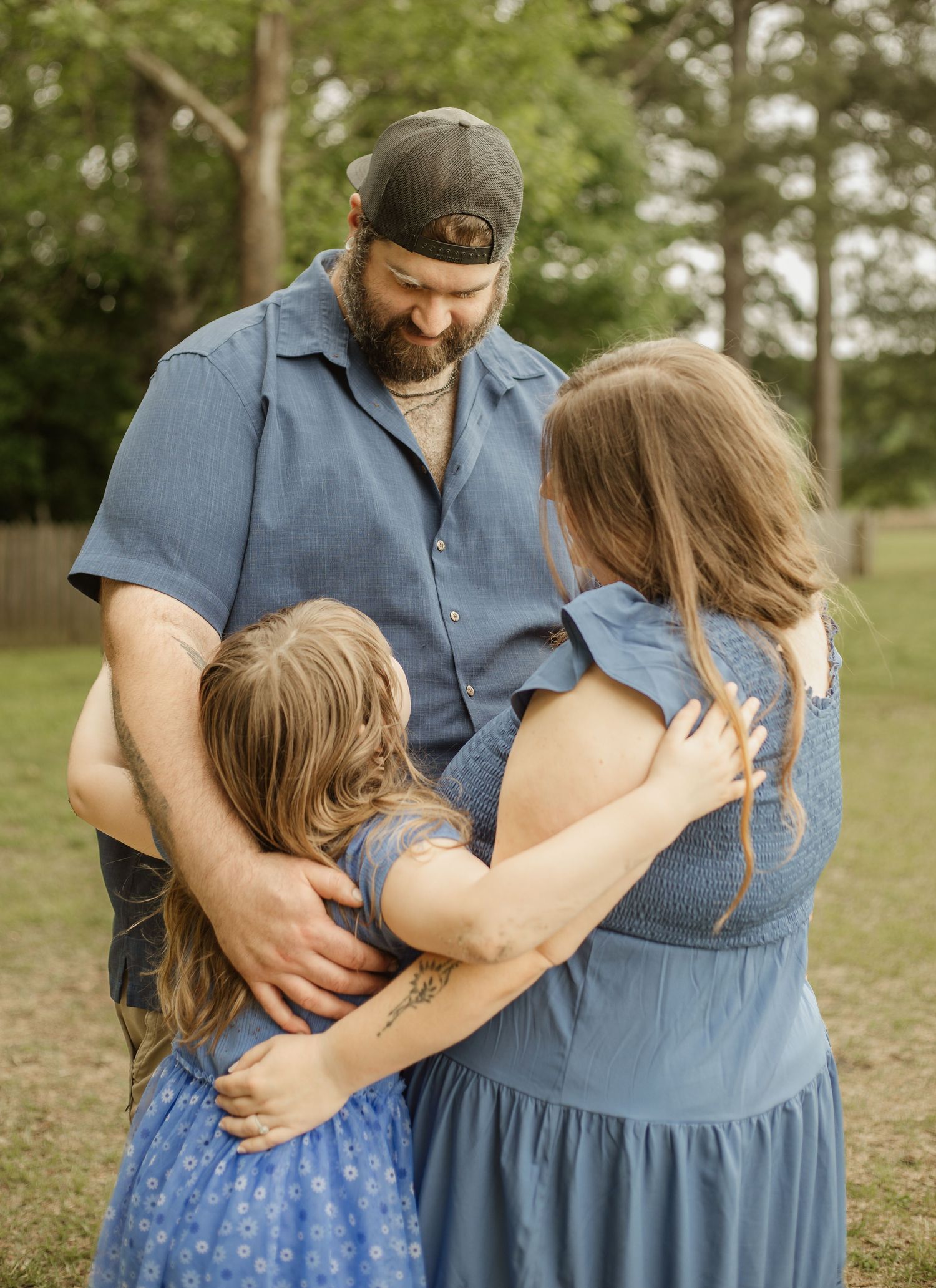 A family of three in matching blue outfits share a warm group hug outdoors among trees.