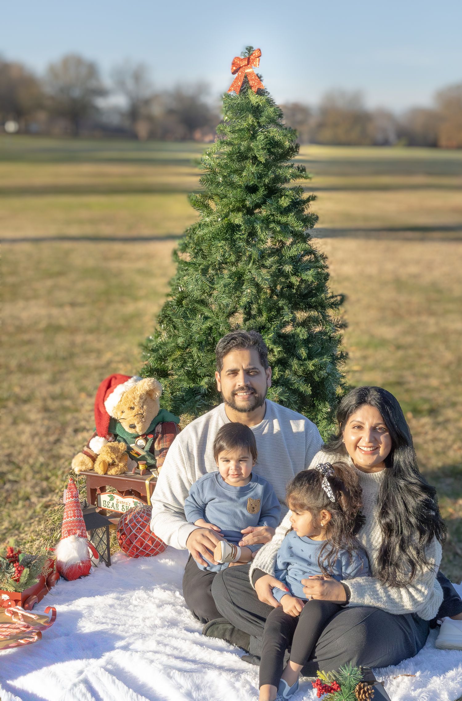 Family sits together on blanket with Christmas tree and holiday decorations outdoors.