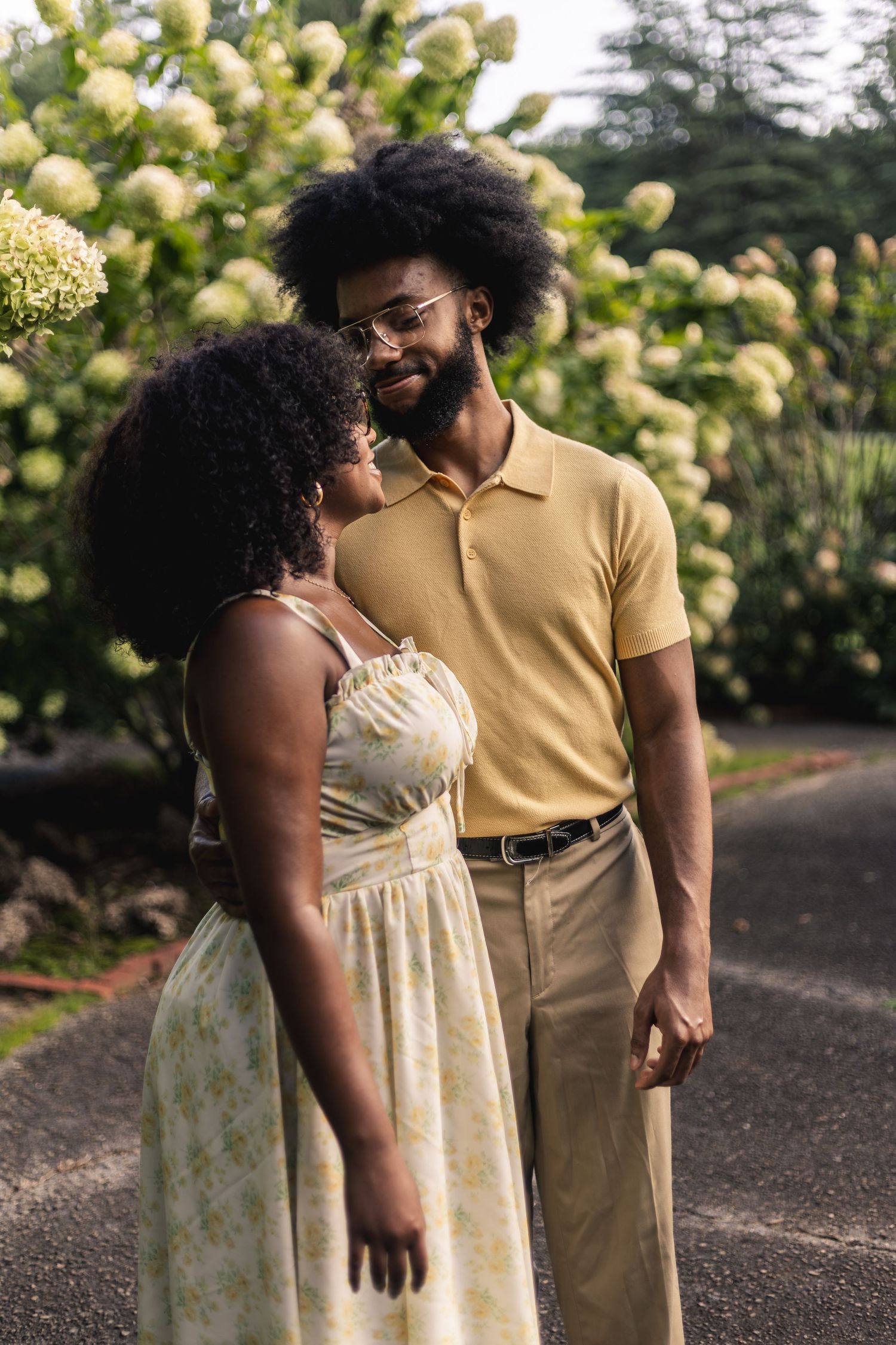 A stylish Black couple in yellow outfits stand together in a lush garden with white hydrangeas blooming behind them.