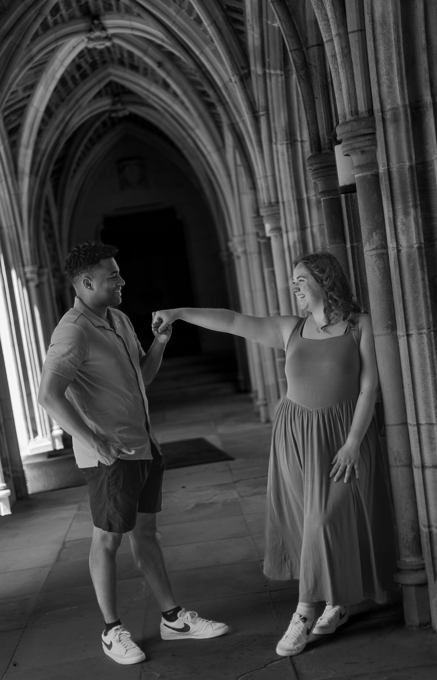 Black and white photo of a couple fist bumping in a gothic stone archway corridor.