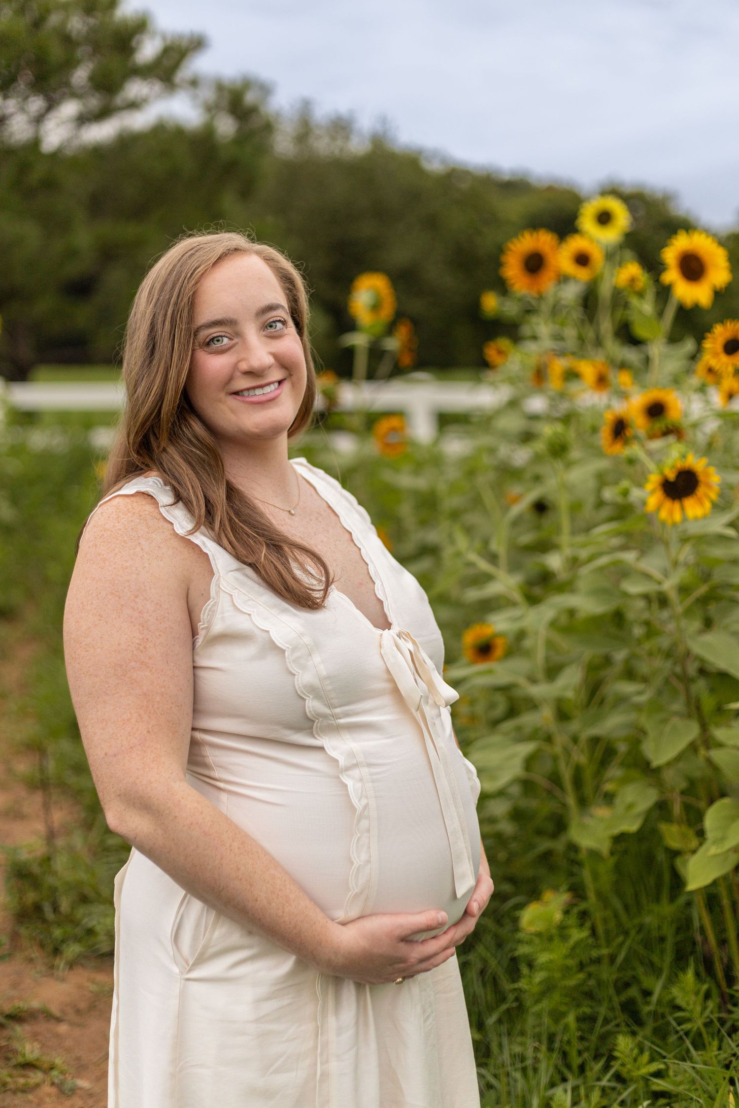 Smiling pregnant woman in white dress holding baby bump beside sunflowers in a garden field.