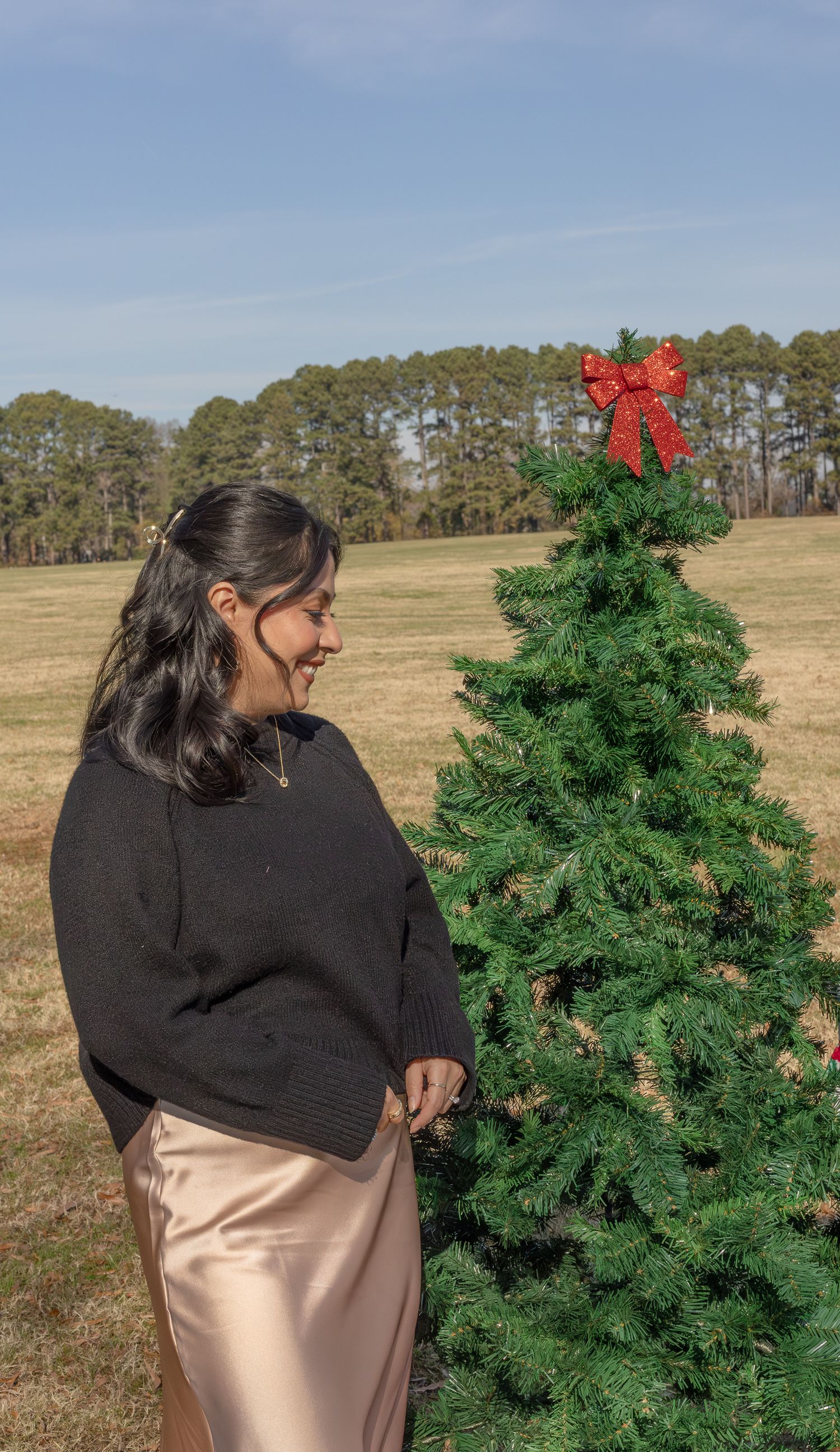 Person in black sweater stands beside decorated Christmas tree in an open field.