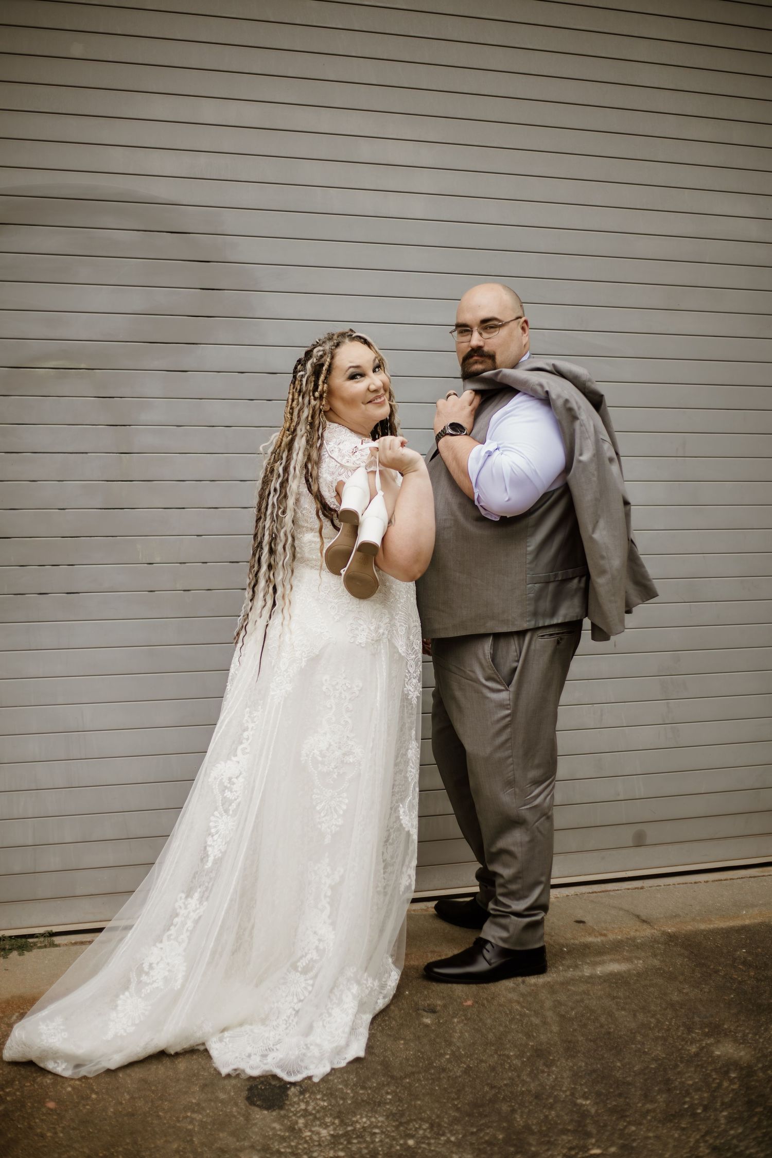 Bride in lace gown with boho braids poses with bald groom in gray suit outside rustic venue.