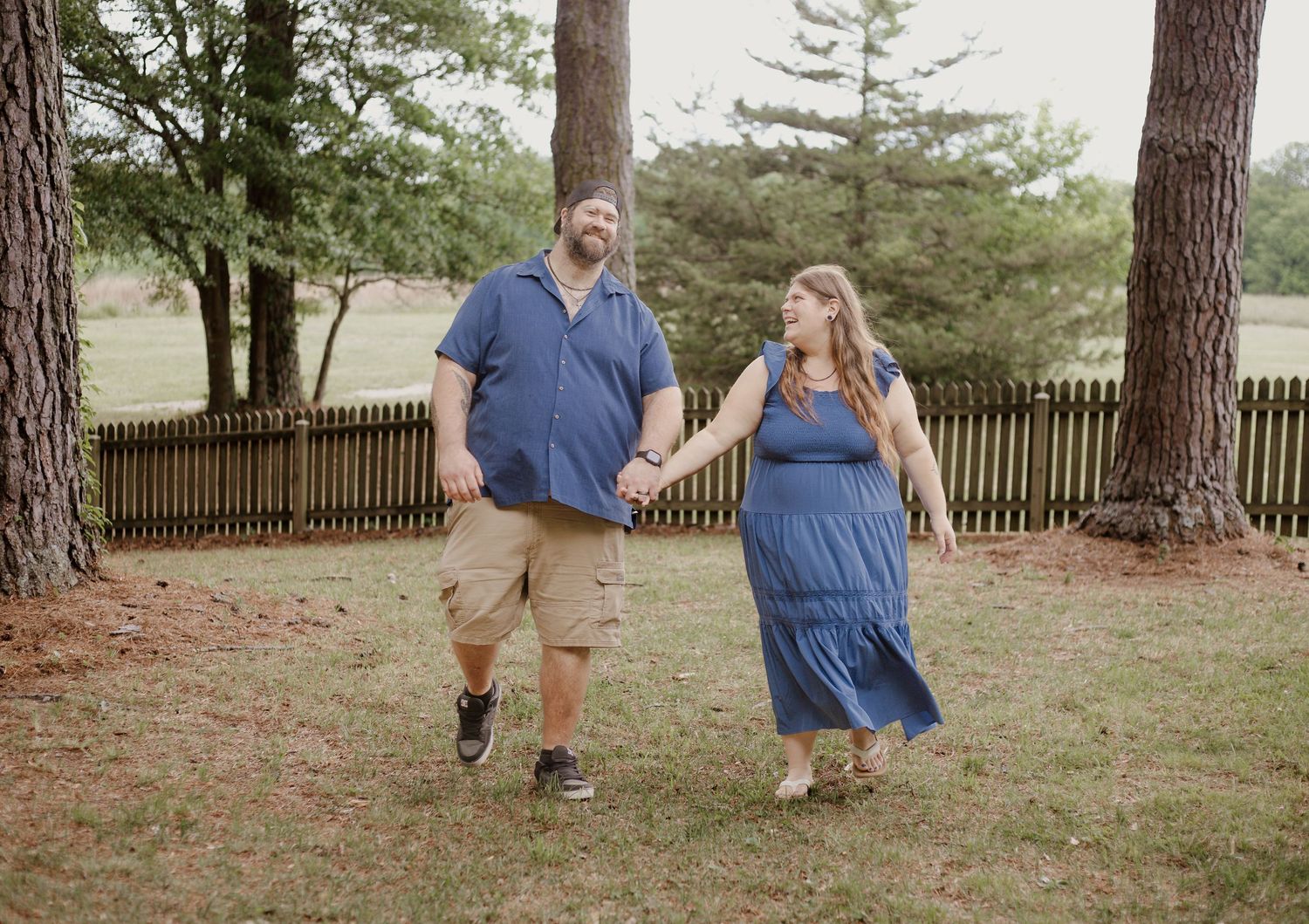Couple holding hands walking outdoors near pine trees with wooden fence backdrop in matching blue outfits.