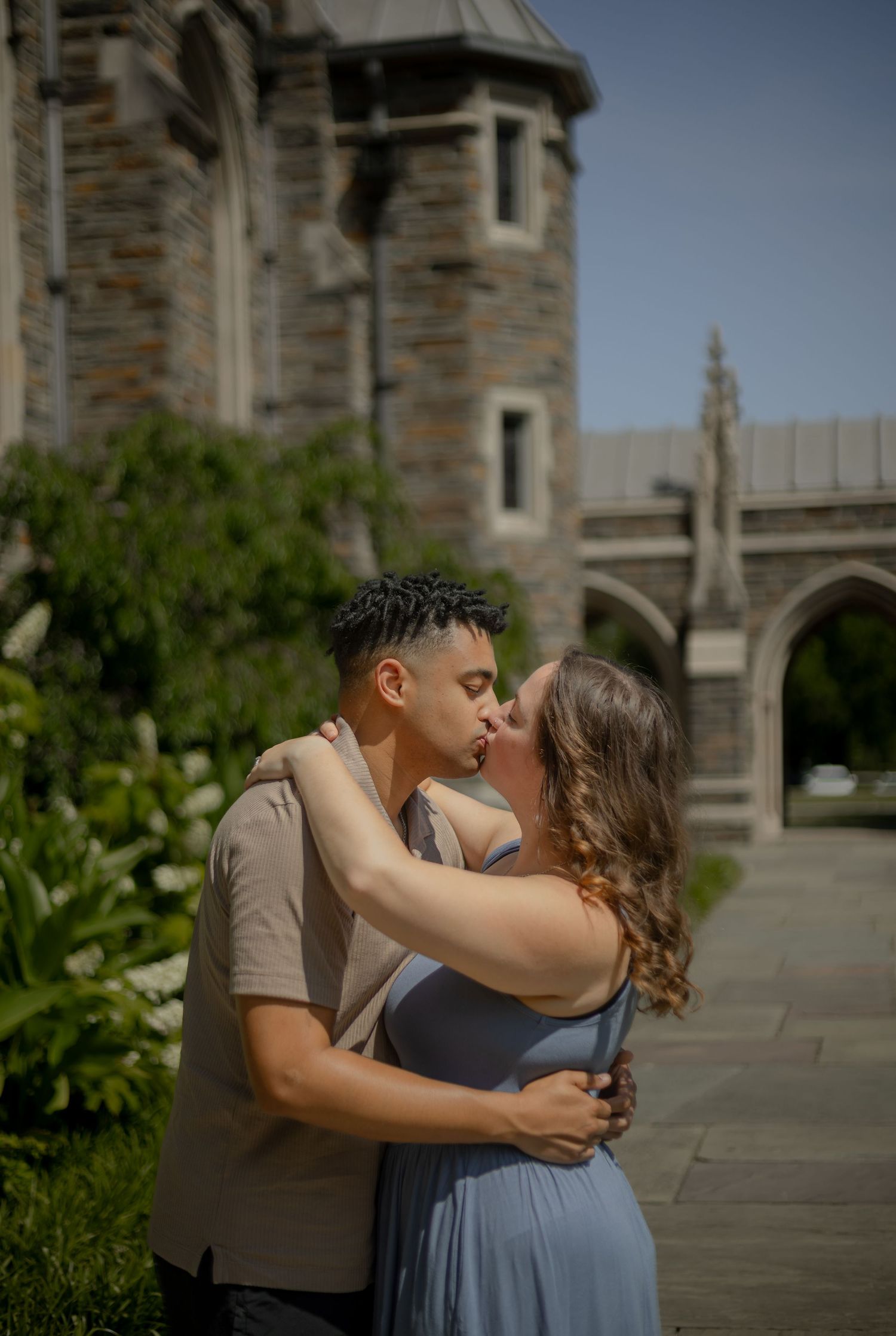Couple sharing a romantic kiss outdoors in front of a historic stone castle building on a sunny day.