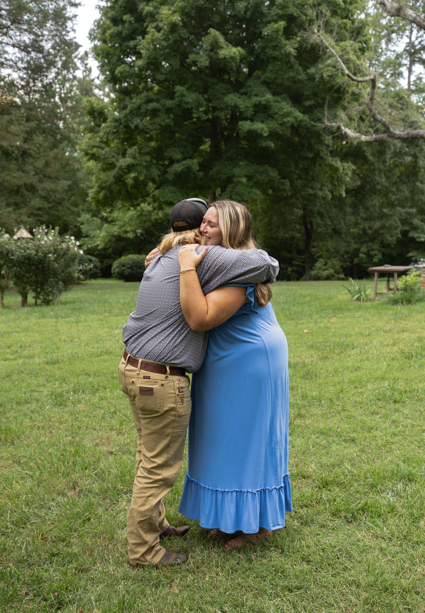 Two people share a warm embrace outdoors on green grass, surrounded by lush trees, one wearing a blue maxi dress.